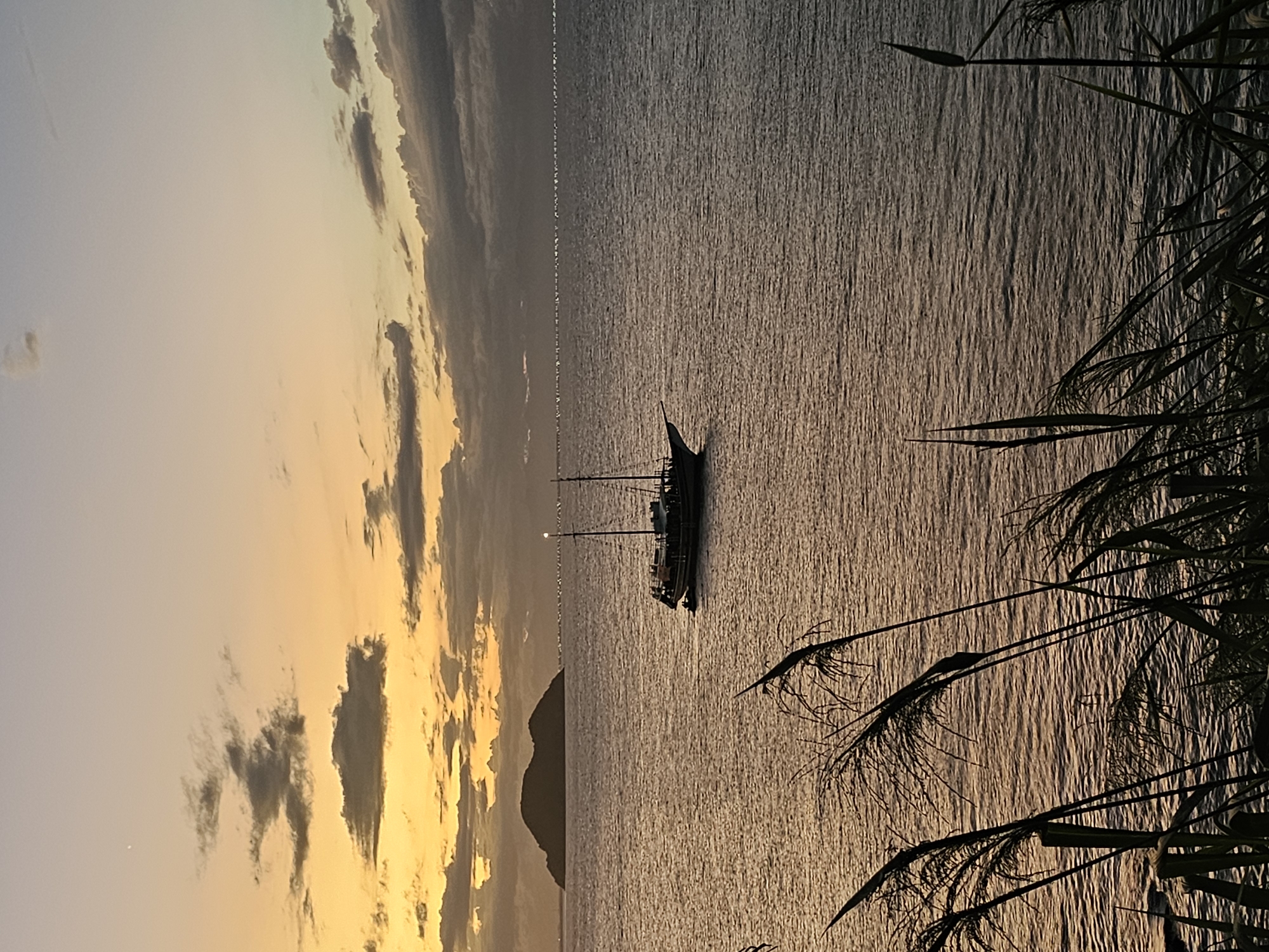 The image shows a coastline in Armação Dos Búzios, Brazil at dusk. A sailboat sits on the water, silhouetted against the setting sun. A low-lying mountain sits near the horizon. Grasses and vegetation frame the scene on the right. Clouds are visible in the golden sky on the left.