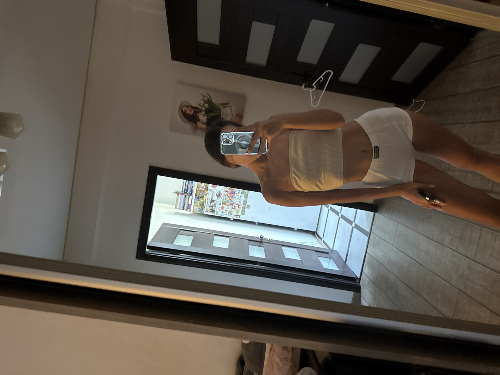 A young woman stands in a bedroom, taking a mirror selfie. She is wearing a white bandeau top and white shorts. In her right hand, she holds a smartphone, which is capturing her reflection. The room features wooden floorboards and a closed door with glass panels. A coat hanger is visible on the door. To the left, a large mirror reflects the scene. Further in the background, through an open doorway, a glimpse of another room with shelves and books can be seen. A framed picture of a woman with flowers hangs on the wall. The overall impression is of a casual, personal space.