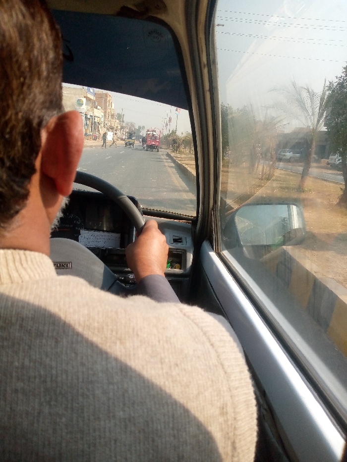 The image captures a driver's perspective from inside a moving vehicle, looking forward through the windshield and to the right through the passenger-side window. The primary subject is the back of a person's head and right shoulder, presumably the driver, seen from behind. They are wearing a light-colored, ribbed sweater and are holding the steering wheel with both hands. Part of their right ear is visible. The dashboard of the vehicle is visible, with the word "SUZUKI" printed on it.
Outside the vehicle, a road stretches into the distance, lined with buildings on the left and trees and concrete barriers on the right. The buildings on the left appear to be shops or commercial establishments, with some people visible walking on the roadside. Further down the road, various vehicles are present, including what looks like a red auto-rickshaw or similar utility vehicle. On the right side, beyond the concrete barrier, there are palm trees and other foliage, with more buildings or structures visible in the hazy distance. A side-view mirror is visible on the passenger door, reflecting the scene to the right.
The scene depicts a typical day on a road in Dijkot, Pakistan, consistent with the architecture, vehicle types, and general environment. The sky is bright but appears somewhat hazy. Sunlight casts a shadow across the driver's back, indicating it is daytime, likely morning or afternoon.