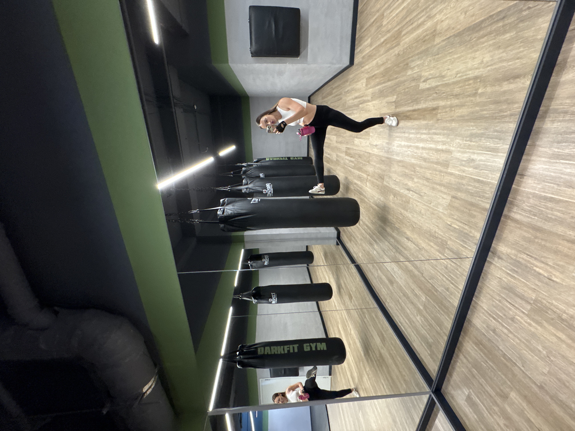 A woman in workout attire takes a selfie in a gym. She is wearing a white sports bra, black leggings, and white sneakers, and is holding a pink water bottle in her left hand. Her right leg is extended behind her, and her body is angled towards the camera as she smiles. The gym features a wooden floor, black punching bags hanging from the ceiling, and mirrors that reflect the scene. The walls are dark blue with a horizontal green stripe. A black square cushion is mounted on the wall. The overall atmosphere appears to be that of a modern, well-equipped fitness facility. The lighting suggests it is daytime, though no windows are visible. The woman appears to be in good spirits, possibly during or after a training session. The text "DARKFIT GYM" is visible on one of the punching bags.