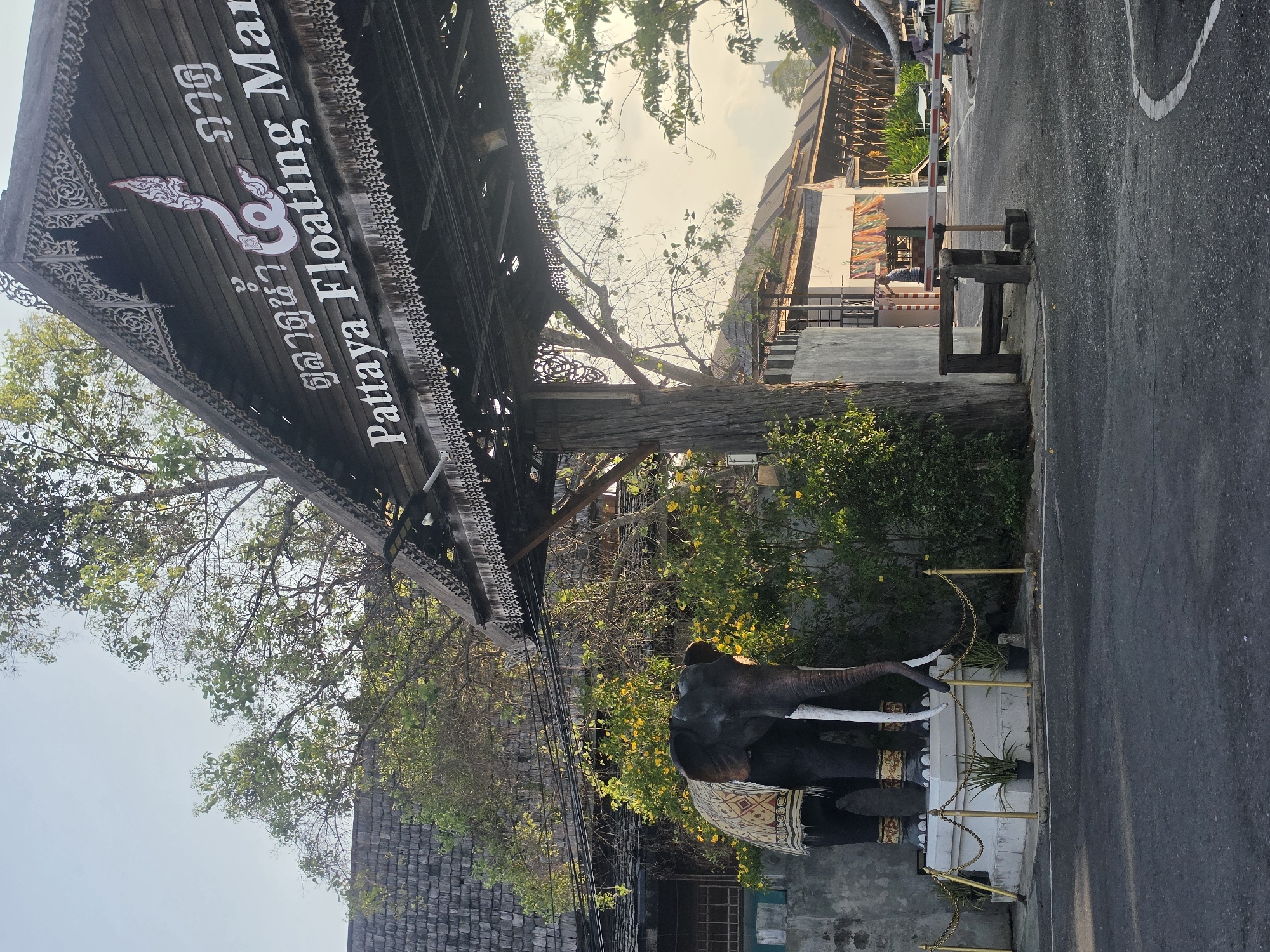 The image captures the entrance to the Pattaya Floating Market in Pattaya City, Thailand, under bright daylight. A prominent, dark wooden arched sign spans the entrance overhead, featuring intricate carvings along its edges. White text on the sign reads "Pattaya Floating Mar" in English, partially visible, and "ตลาดน้ำ ๔ ภาค" (Talad Nam See Pak), which translates to "Four Regions Floating Market" in Thai. A stylized, serpent-like symbol, likely a Naga, is centrally integrated into the sign's design.
To the right of the entrance, positioned beside a paved asphalt road, stands a large, dark elephant statue with white tusks and an ornate patterned blanket draped over its back. The statue is enclosed by a low golden chain barrier, with small potted plants placed along its base. Abundant green foliage and trees with scattered yellow blossoms frame the scene, creating a natural backdrop. In the background, beyond the entrance, traditional-style wooden structures are visible, along with a white and red barrier arm. A single person can be faintly seen near this barrier. The clear sky and strong sunlight indicate a sunny day, suggesting the welcoming ambiance of a tourist destination.