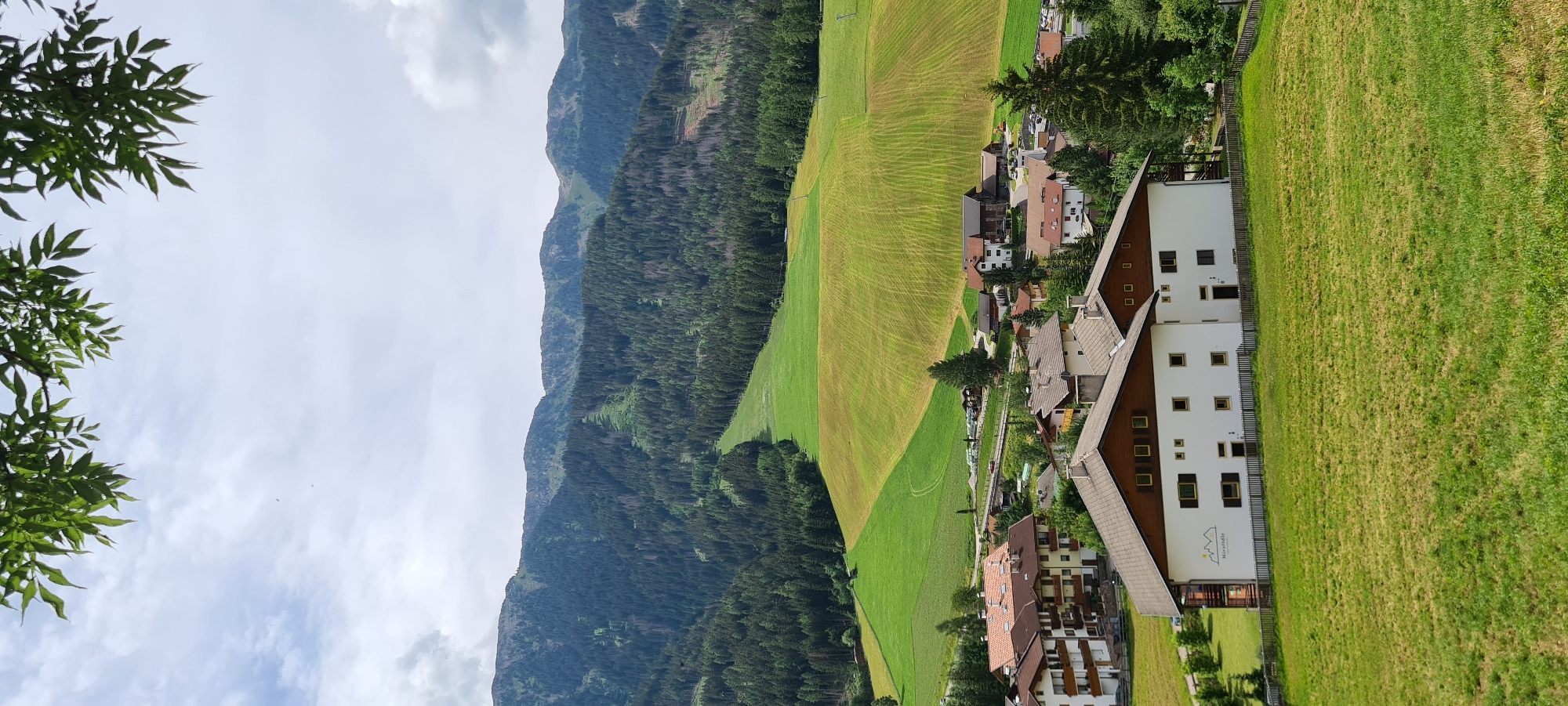 This is a vertical, eye-level, full shot of a village nestled in the Italian Alps. The foreground features a lush green meadow sloping upwards. In the middle ground, a cluster of buildings with traditional alpine architecture, characterized by wooden trim and brown tiled roofs, are visible. One prominent white building in the foreground has a logo that appears to read "Mirabello." Beyond the village, rolling hills transition into a dense forest of evergreen trees, which climbs towards the majestic, rocky mountains that dominate the background. The sky above is a mix of white and grey clouds, suggesting a dynamic weather pattern. There are no people or animals visible in the scene. The village is situated in the municipality of Funes, South Tyrol, in the geographical region of St. Magdalena - Santa Maddalena, Italy, famous for its picturesque scenery.