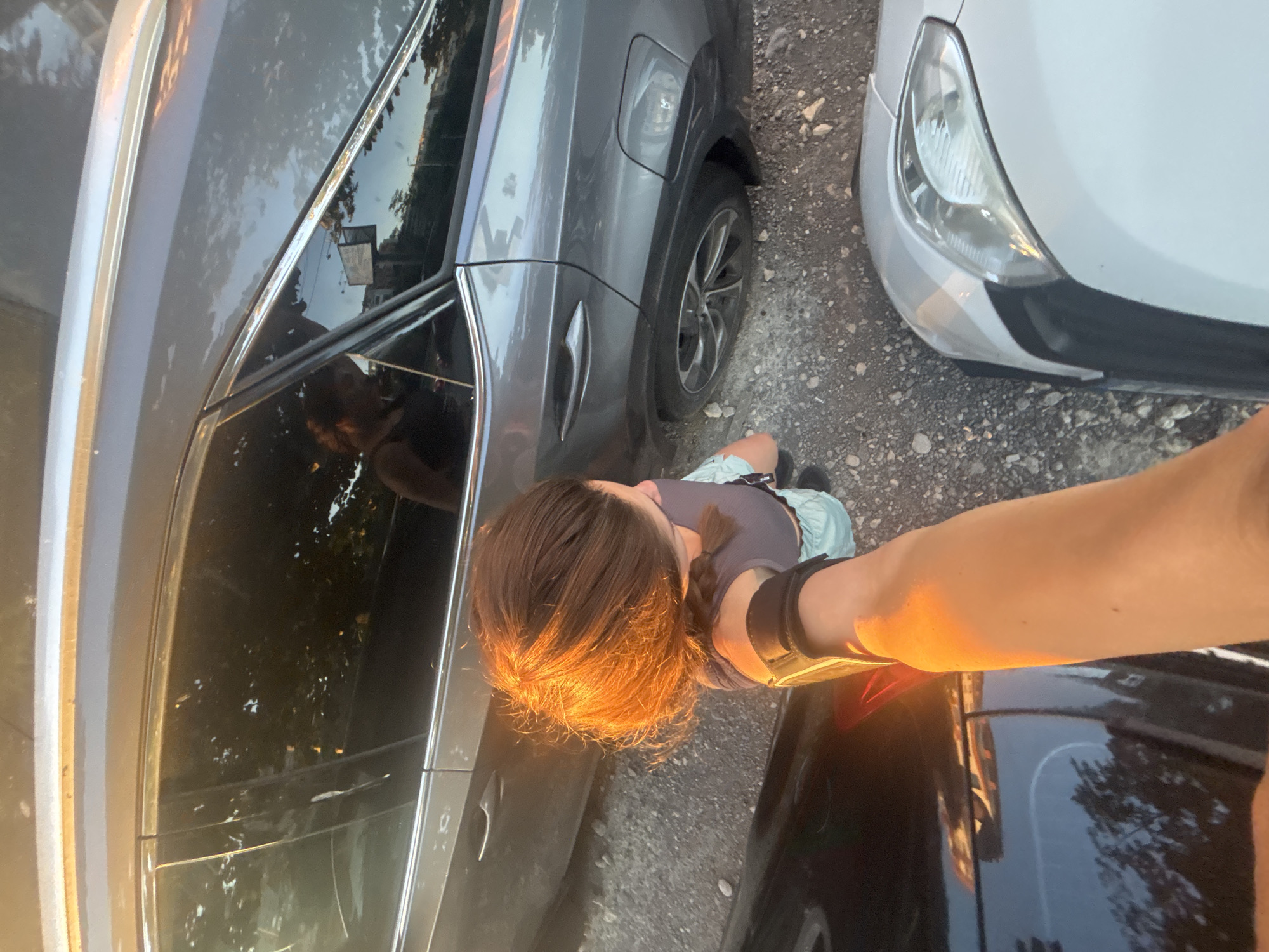 The image is a first-person perspective of a woman standing between two parked cars, with a third car partially visible in the foreground. The woman, with her hair in a braid and wearing a grey top and light blue shorts, is looking down and to the side. Her left arm, extended forward and holding a device, is prominently featured in the lower part of the image, illuminated by golden sunlight.
The cars appear to be in a parking lot or on a street with a gravelly surface. The car to her right is a dark grey SUV, and the car to her left is black. A white car is partially visible in the foreground. The windows of the grey SUV reflect trees and the sky, hinting at an outdoor setting, possibly during daylight with the sun low on the horizon due to the warm lighting. The image does not contain any specific landmarks or features that would allow for precise localization within Varna, Bulgaria.