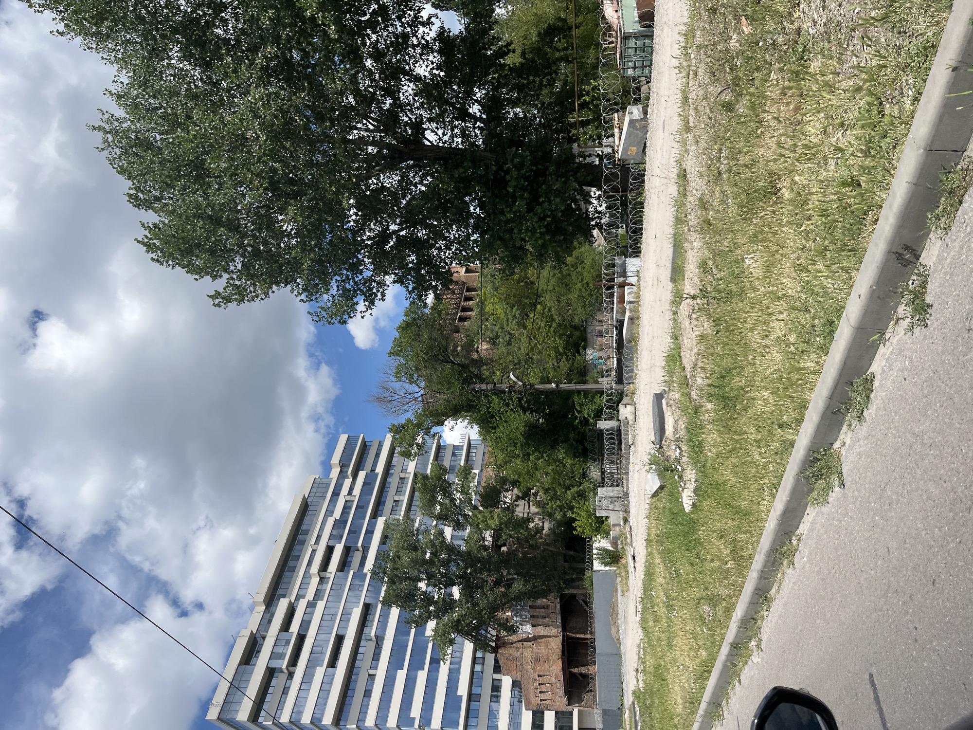 The image depicts a view from a street in Dnipro, Ukraine, showing a juxtaposition of old and new architecture amidst an urban landscape. In the foreground, a paved road curves away, bordered by a concrete curb and an area of overgrown grass and weeds. A black car mirror is visible on the left edge of the frame.
Dominating the mid-ground and background are several structures. To the left, a modern, multi-story building with a facade of white horizontal elements and reflective blue-tinted windows stands prominently. Adjacent to it, to the right, are the decaying ruins of a brick building, its walls partially collapsed and showing exposed internal structure. Several mature trees with dense green foliage are interspersed between these buildings and in the background, suggesting a mix of developed and semi-wild areas.
Further back, a large, mature tree with a thick trunk and expansive canopy is a significant feature on the right side of the image. Behind the trees and ruins, hints of other structures and greenery are visible. A wire, likely a power or communication line, stretches diagonally across the upper portion of the image against a sky filled with white cumulus clouds.
The ground in the mid-ground area appears to be a mix of dirt, gravel, and sparse vegetation. Barbed wire fencing is visible in several places, indicating a boundary or security measure around some of the areas, particularly around the ruins and what appears to be a collection of construction materials or debris. Concrete blocks and other urban detritus are scattered across the ground. There are no clearly discernible people or active engagements in the scene, presenting a static snapshot of an urban environment in a state of transition or disrepair alongside modern development.