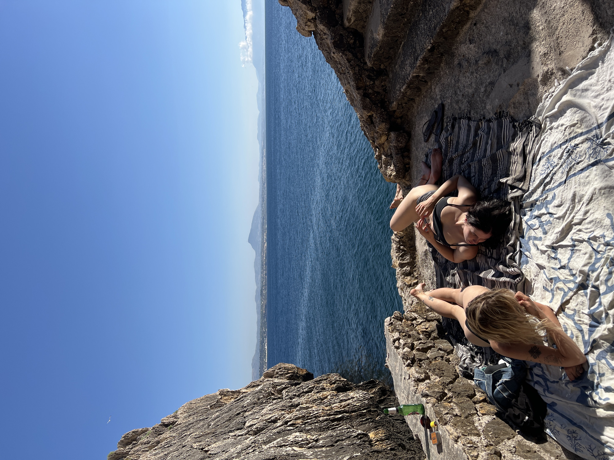 Two women are sunbathing on a rocky cliff overlooking a body of water. The woman in the foreground has blonde hair and is lying on a patterned blanket, facing away from the camera. She is wearing a black bikini and has tattoos on her arms and legs. The woman behind her has dark hair and is lying on a striped blanket, also in a black bikini. Both appear relaxed and are enjoying the sunny weather.
The scene is set outdoors on a cliffside with rough, rocky terrain. A blue towel with a white and blue pattern is spread out next to the striped blanket. A pair of dark flip-flops are on the striped blanket. There are also some clothes, possibly denim shorts, on the ground. A green glass bottle, possibly a beer bottle, and a small yellow object are also visible on the rocks.
The sky is clear and blue, indicating a sunny day. The sea is a deep blue and appears calm. In the distance, a coastline with hills and buildings can be seen, suggesting a populated area. The overall mood is one of relaxation and enjoyment of a summer day by the sea. The time of day appears to be daytime, likely late morning or early afternoon, given the bright sunlight casting shadows. The location is identified as Terrasini, Italy. No visible text is present in the image.