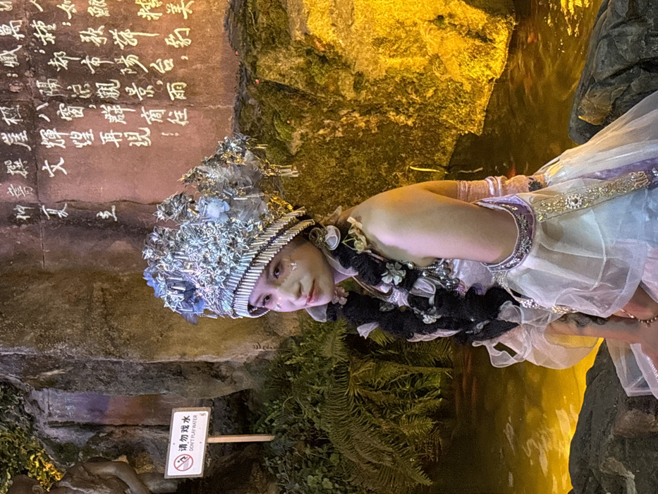 A young female individual is seated and looking towards the viewer in an artificially lit outdoor environment, specifically in Yuzhong District, China. She is adorned in an elaborate costume, featuring a large, ornate silver-colored headdress with intricate designs and light blue accents. Her dark hair is styled into two thick braids, decorated with small, light-colored floral elements, extending over her shoulders. Her face is made up with glitter-like embellishments on her cheeks and around her eyes. She wears a multi-layered gown in shades of white, pink, and purple, with sheer fabric overlays, metallic-embroidered elements, and decorative armbands. She is positioned on dark rocks at the edge of a body of water that reflects a warm, yellow-orange light source. In the background, a textured rock wall is visible. The section to the right is brightly illuminated with yellow light and features white Chinese calligraphy. The section to the left is darker, covered with green fern-like foliage. A white rectangular sign with a red circular prohibition symbol is present on the left, bearing the text "请勿戏水 / DON'T PLAY WATER" on a wooden post. A partial view of a light-colored stone sculpture is visible on the far left, behind the sign and foliage.