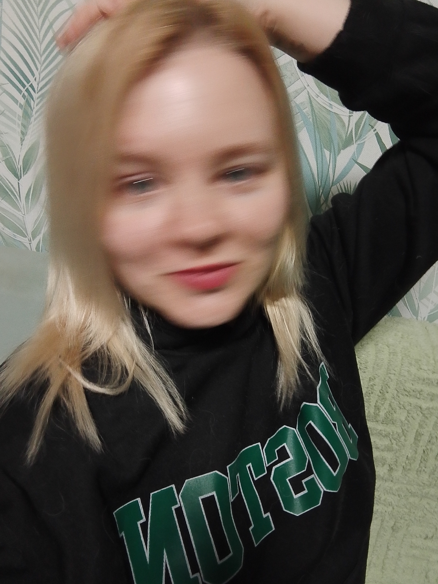 A blurry selfie shows a woman with blonde hair wearing a black sweatshirt with the word "BOSTON" in green and white lettering. Her right hand is raised to her head. The background features wallpaper with a green leaf pattern and a green textured cushion. The location cannot be determined as Izhevsk, Russia from the image.