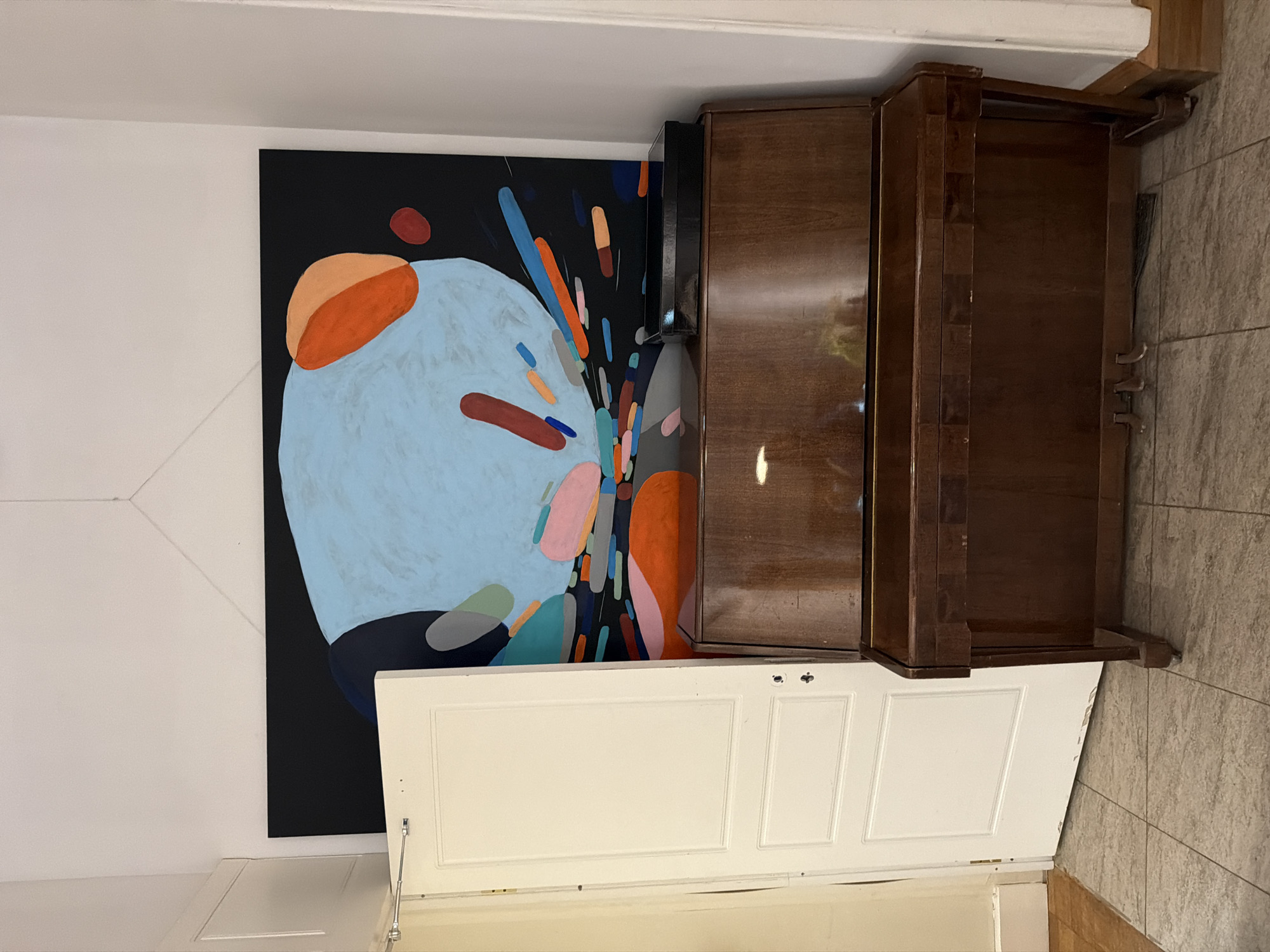 The image shows an interior space with a painting, a cabinet, and a door. The painting features abstract shapes and colors on a black background. The cabinet is dark wood and sits near the painting. A white door is positioned in front of the painting and cabinet. The location is uncertain, but the image appears to be taken indoors. There is no evidence of Moscow, Russia.