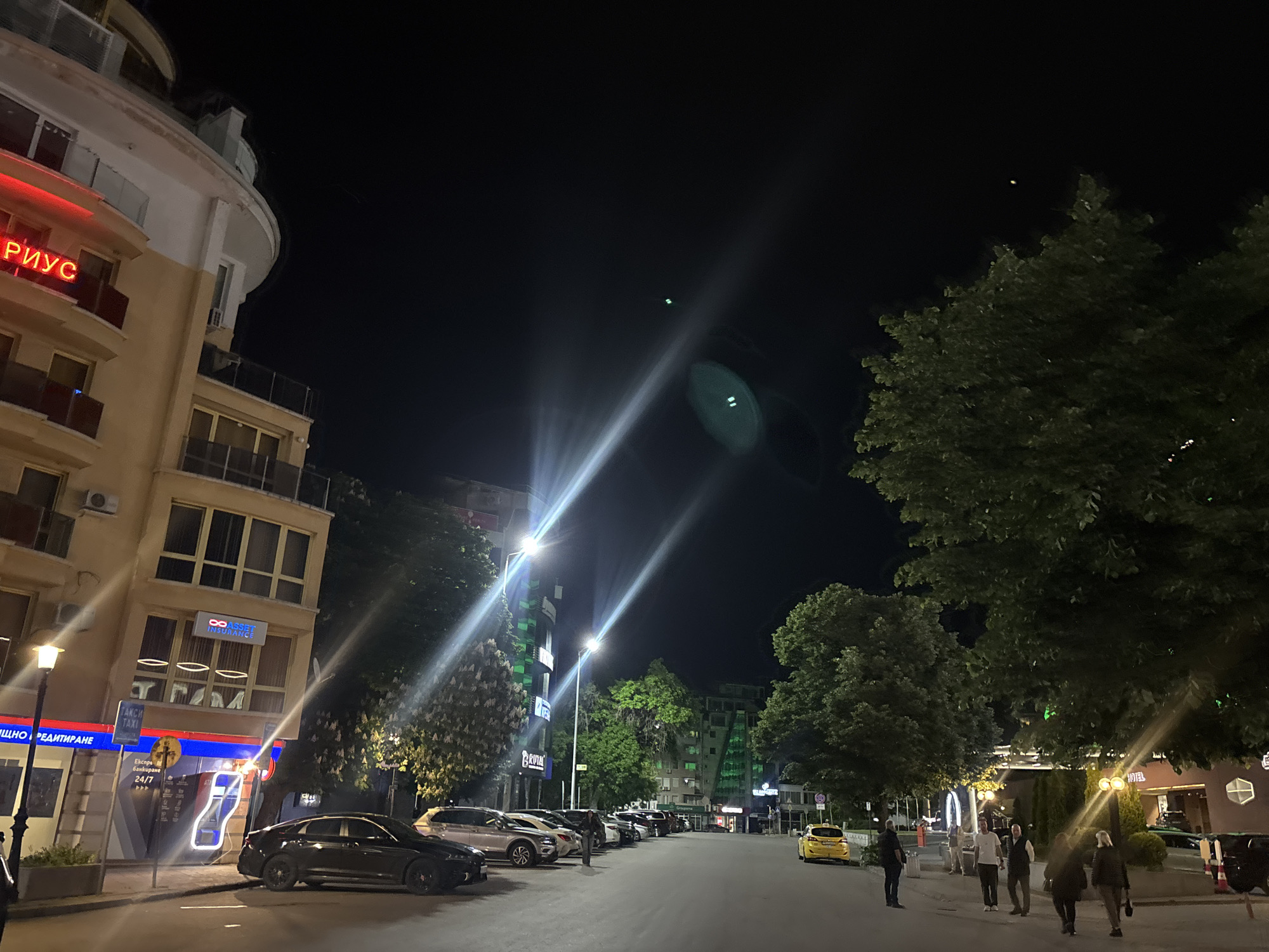 A night scene depicts a street in Plovdiv, Bulgaria. A beige building with a rounded corner and red neon signage stands on the left. Along the street are parked cars and interspersed trees. Pedestrians are seen walking in the direction away from the camera. Street lamps provide illumination. The sky is dark. There are shops and businesses visible along the street.