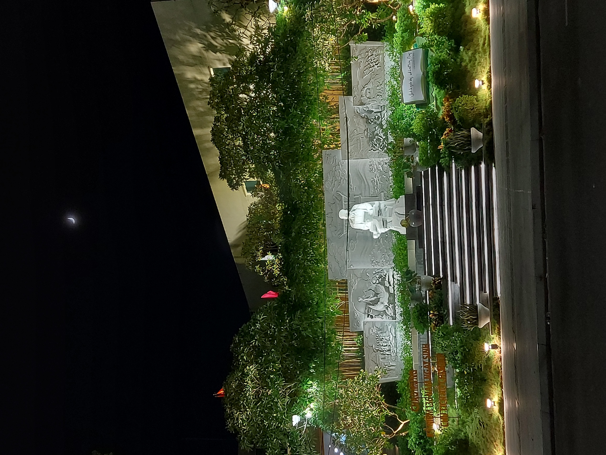 The image depicts a nighttime scene in Da Nang, Vietnam, featuring a monument with carved reliefs and a central statue. The monument is illuminated by spotlights, highlighting its intricate details. Lush green trees and plants surround the monument, creating a vibrant contrast with the dark night sky. A crescent moon is visible in the upper portion of the sky, adding to the atmospheric quality of the scene. The overall impression is one of solemnity and commemoration, with the lighting and natural elements contributing to a sense of tranquility.