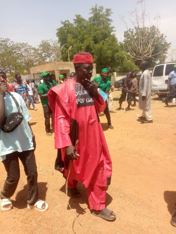 The scene depicts an outdoor gathering in Kagoma, Nigeria, under bright daylight with a clear sky. The ground is unpaved, appearing dry and reddish-brown.
In the foreground, a dark-skinned man is centrally positioned, facing slightly to his right. He wears a red traditional cap and a long, flowing red garment over a black undershirt. The black undershirt features white text that reads "DOGARI POP GWG". He has a short, dark beard and his right hand is raised, with fingers touching his chin. His left hand holds a thin, light-colored rope or cord. He wears dark sandals. To his immediate left, partially visible, is a younger individual wearing a light blue t-shirt, dark trousers, white sandals, and a black shoulder bag. This individual faces left, away from the central figure.
In the mid-ground, several individuals are present. A group of men, many wearing green shirts and green caps, are visible behind the central figure. One man within this green-clad group appears to be holding a shiny, brass wind instrument, possibly a trumpet or bugle, partially obscured. Further to the right of the central figure, another man in a light-colored (possibly off-white or light grey) traditional tunic and trousers stands, facing left. In the deeper mid-ground, another individual is seen holding a long stick or staff.
The background features a light-colored, possibly concrete or mud brick building with a flat roof, located to the left. Various green trees and sparse vegetation are scattered across the background, including a prominent tree with dense green foliage to the left and a bare, branchy tree to the right. A white passenger van is parked or moving on the far right side of the frame. Shadows are visible on the ground, indicating a high sun angle.