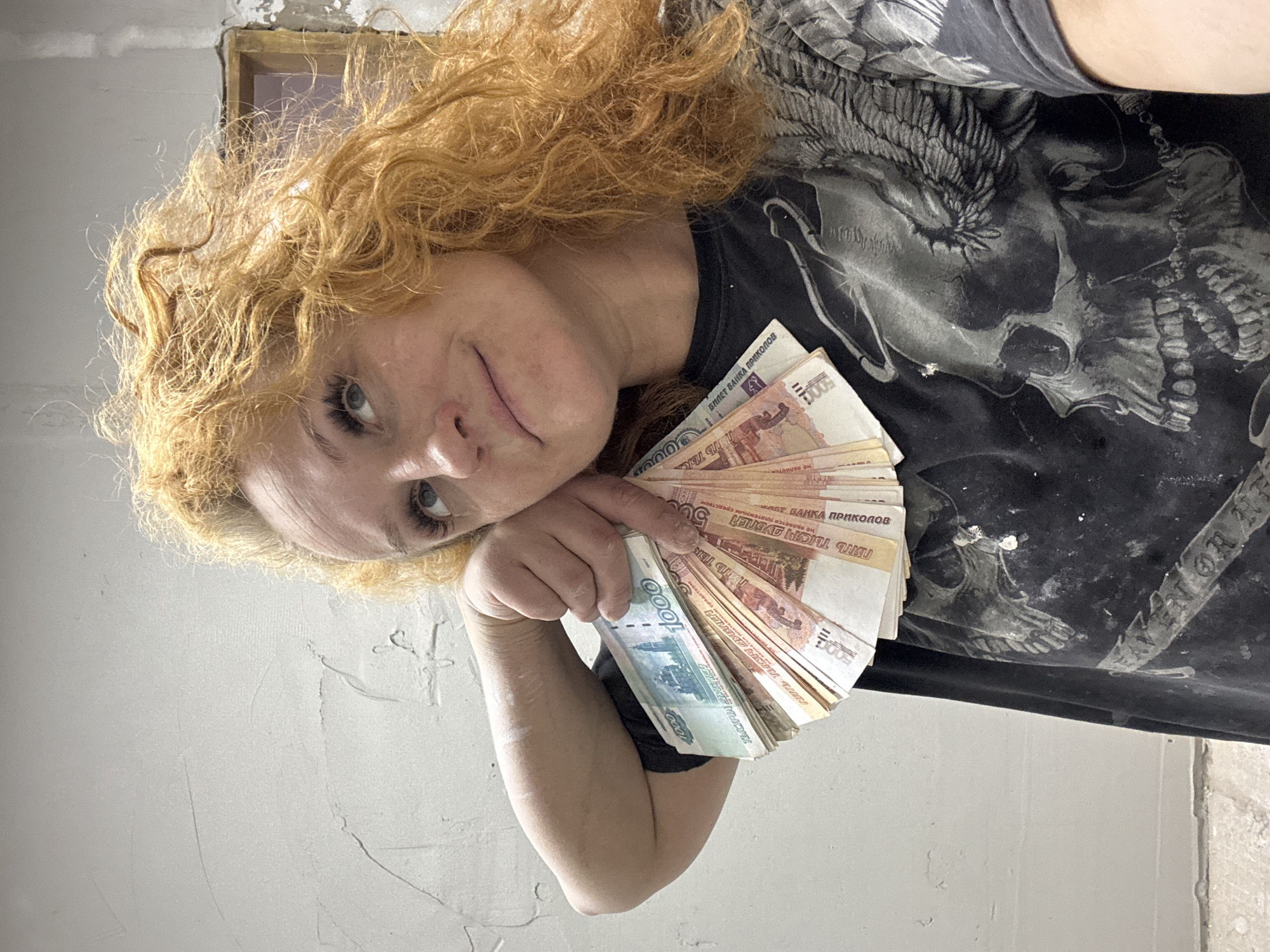 The image, taken in Chaikovsky, Russia, features a person, likely a woman, with voluminous, curly, reddish-blonde hair, looking directly at the camera. She is captured from a low-angle perspective, giving the appearance of gazing down at the viewer. She wears a dark t-shirt adorned with a large grey and white design depicting a skull and wings; partially visible text on the shirt reads "AVEN OR HE".
In her right hand, held across her chest, she displays a fan of banknotes. These are Russian Ruble denominations, specifically 1000 and 5000 Ruble notes. A significant detail is that many of the 5000 Ruble notes are clearly marked as novelty items, bearing inscriptions like "БИЛЕТ БАНКА ПРИКОЛОВ" (Joke Banknote) and "НЕ ЯВЛЯЕТСЯ ПЛАТЁЖНЫМ СРЕДСТВОМ" (Not a means of payment). However, the 1000 Ruble notes appear to be genuine currency.
The person has light blue eyes and a subtle, almost smirking expression. The background consists of plain, light-colored walls, possibly unpainted or roughly plastered, indicating an indoor, unadorned, or potentially unfinished setting. A small, dark, rectangular opening or frame is visible in the upper background. The overall scene is informal and direct.