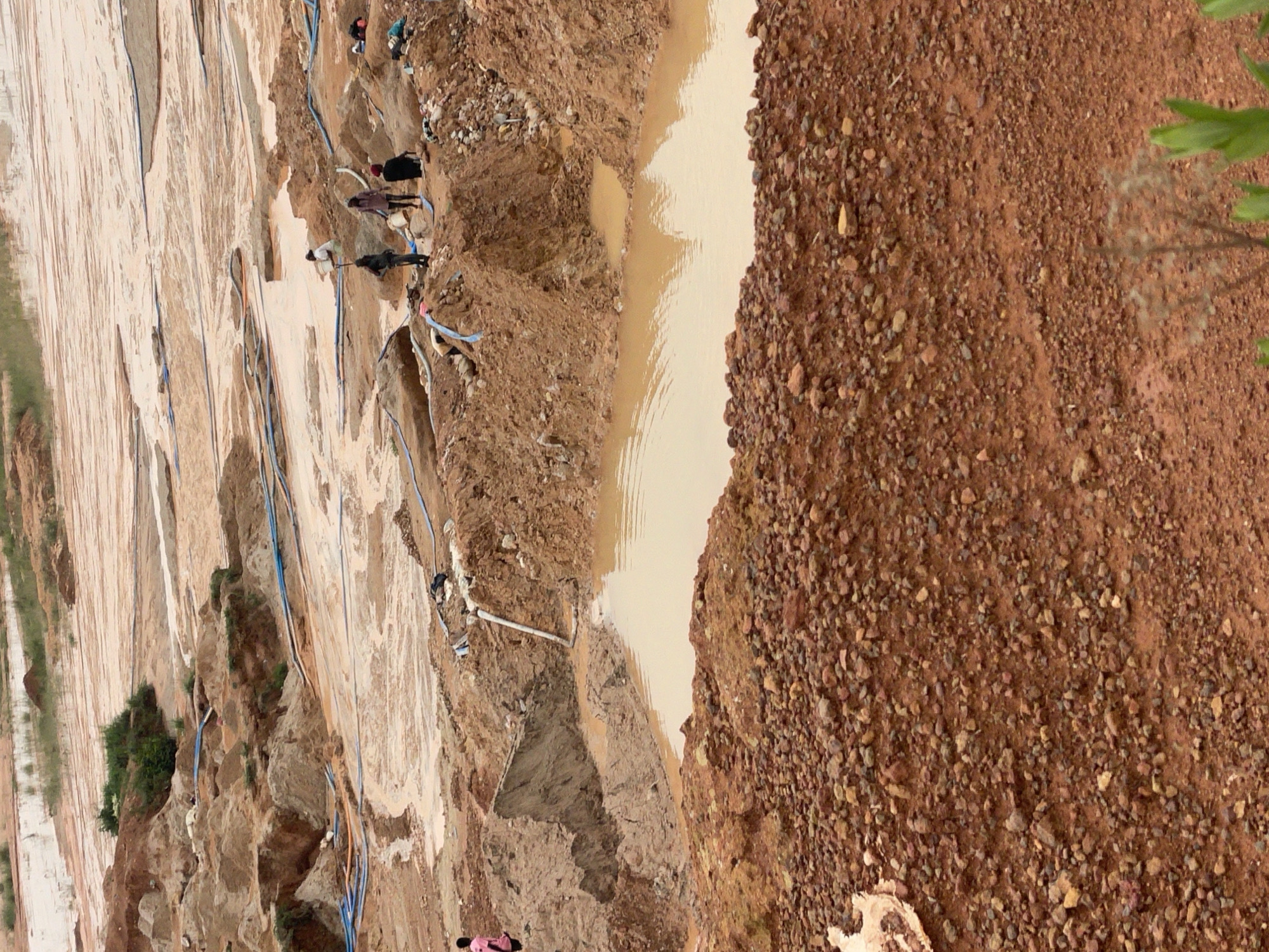 The image depicts a wide shot of a large, open-pit mine in Jos, Nigeria. The landscape is characterized by its reddish-brown earth, with visible layers of soil and sand that have been excavated. Several water features, appearing as muddy pools and streams, are scattered throughout the scene, likely a result of recent rainfall or mining operations. Blue and orange pipes are intermittently laid across the ground, suggesting a system for water or material transport.
Several individuals are present at various locations within the mine. In the foreground, on the right side of the image, a person wearing a pink shirt stands near a large pile of reddish-brown rocks and soil. Further into the mid-ground, a group of individuals is clustered together. One person, wearing a white top and dark trousers, appears to be working with a tool. Others in this group are dressed in various casual clothing. In the background, more people are visible, along with additional water-filled depressions and excavated areas. The overall impression is one of active mining, with the environment shaped by the extraction process.