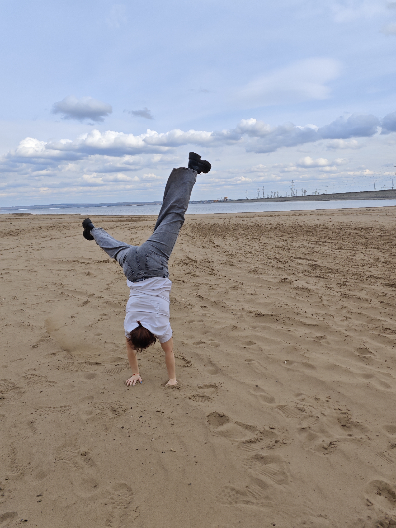 A person is doing a handstand on a sandy beach. The person is wearing a white t-shirt, gray jeans, and black shoes. The beach is near water, and in the distance, there are power lines and industrial structures. The sky is cloudy. The location is near Noviy, Russia.