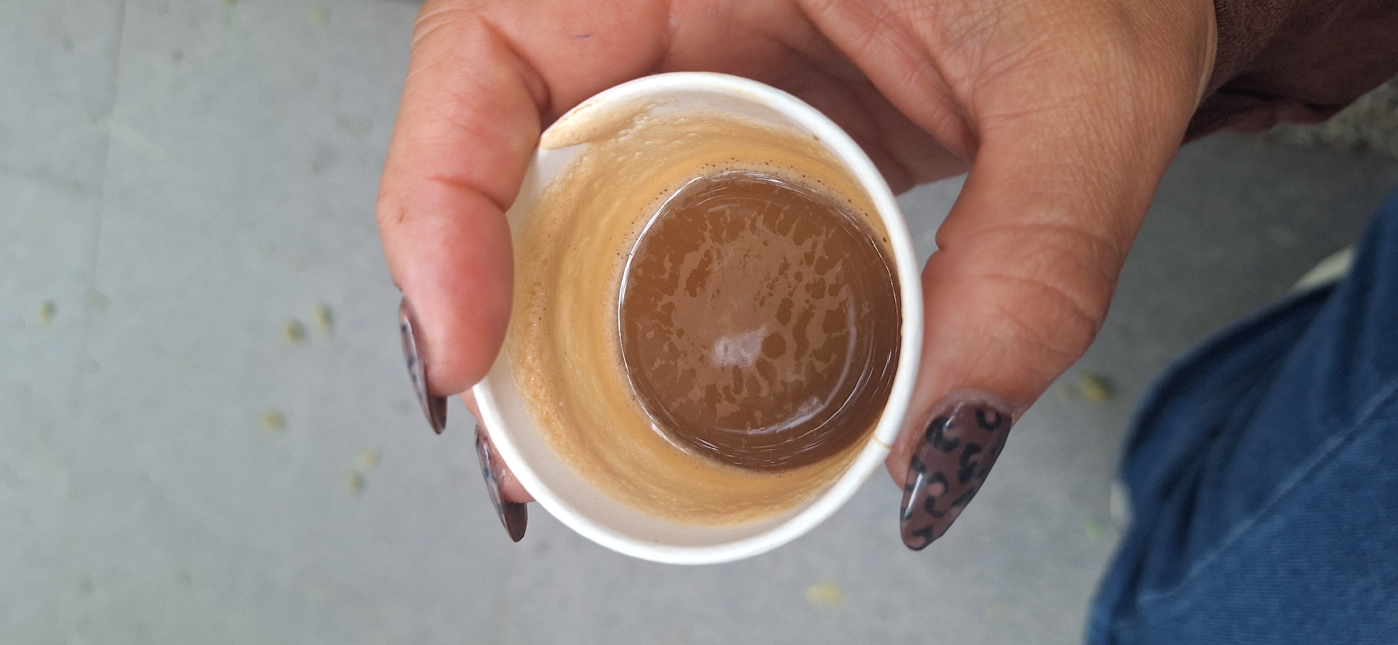 An overhead close-up shot captures a person's hand holding a small, white disposable cup filled with coffee. The hand exhibits a medium skin tone, and the fingernails are almond-shaped, adorned with a distinctive dark brown and black leopard print design. The cup contains a rich, dark brown liquid, indicative of espresso or strong coffee, with a prominent layer of light brown crema and small bubbles visible on its surface. The liquid occupies roughly half to two-thirds of the cup's volume.
The background is softly blurred, presenting a light gray, textured surface, possibly concrete or pavement, scattered with indistinct yellowish-green specks. This suggests an outdoor or casual urban setting, consistent with common scenes in Santa Cruz de Tenerife, Spain. A small section of blue fabric, likely denim jeans, is visible in the lower right corner, indicating the person may be seated. The image portrays a simple, casual moment, possibly a coffee break. No text is visible within the frame.