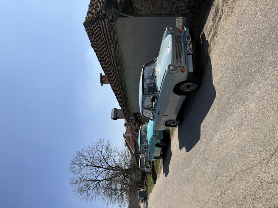 The image displays a street scene in Bukovets, Bulgaria, under a clear, bright blue sky. A cracked asphalt road extends into the distance, with a row of five vintage automobiles, primarily Trabant 601 models, parked parallel to the right curb.
The nearest vehicle is a light blue Trabant 601 sedan with a Bulgarian license plate "CA 2922 TM" on its front bumper. Behind it is a bright turquoise Trabant 601 sedan. The third car in the line is a white Trabant 601 sedan, which appears to have its hood open or removed, exposing engine components. Further down the road, two additional vintage vehicles are partially visible, one of which appears yellow or orange.
To the right of the parked cars, several modest buildings with light-colored, stucco-like walls and terracotta tile roofs line the street. Multiple chimneys are visible on the rooftops. On the left side of the road, a large, leafless deciduous tree stands prominently, indicating a late winter or early spring season. Strong sunlight illuminates the scene, casting distinct shadows from the vehicles and structures onto the road surface. No individuals are present in the image.