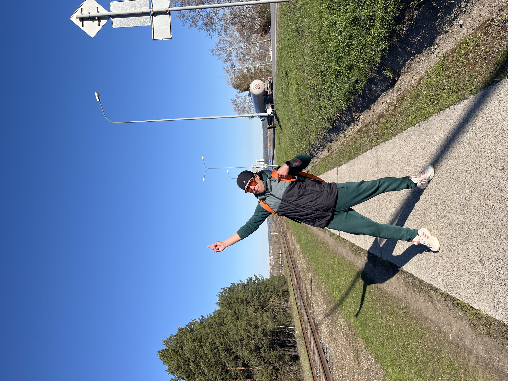 A man wearing a black vest, dark green tracksuit, black cap, and orange-tinted sunglasses stands on a concrete walkway. He is pointing to the left of the frame. Behind him are railroad tracks, grass, and trees. In the background, a tanker truck is parked along a road. A street light and road signs are also visible. The sky is clear and blue. The image was taken in the city of Chaikovsky, Russia.