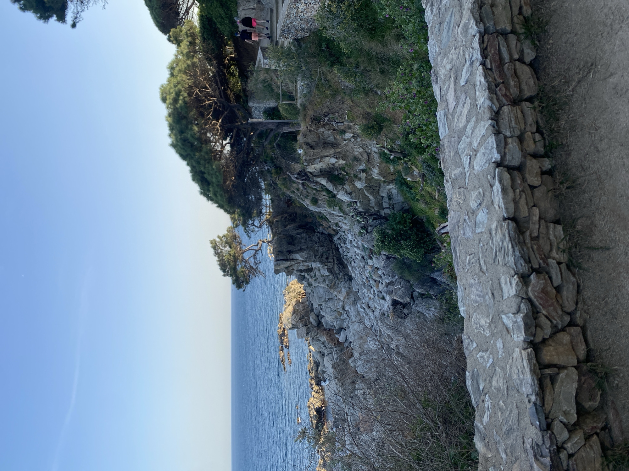 The image captures a scenic view of Calella De Palafrugell, Spain. The foreground showcases a stone wall bordering a walking path. Beyond the wall, the land drops to a rocky cliff face descending towards the sea. A few trees, some clinging precariously to the cliff, are visible. In the upper part of the image, two people can be seen standing in the distance. The calm, blue water of the sea extends towards the horizon, meeting a clear, pale blue sky.