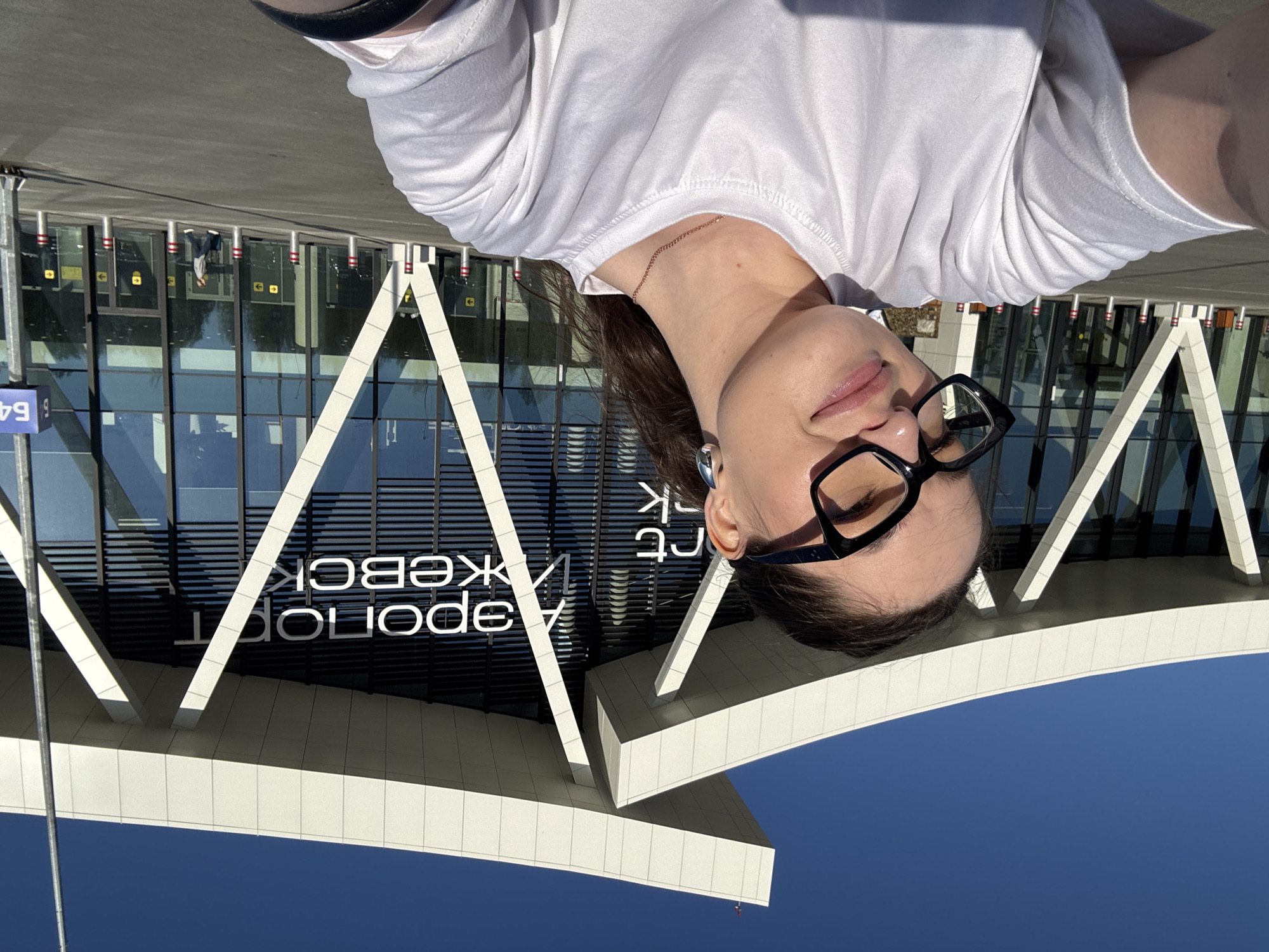 A selfie photograph shows a woman wearing glasses and a white T-shirt in front of a building. The woman is positioned in the upper portion of the frame, her face partially visible, with a silver earring. In the background is a building with black-framed windows and white architectural supports. The facade has the words "Аэропорт Ижевск" (Airport Izhevsk) written on it. A blue sign with the number "64" is visible on the left. The sky is bright blue. The location is identified by the text on the building as Izhevsk Airport, located near Новое Мартьяново, Russia.