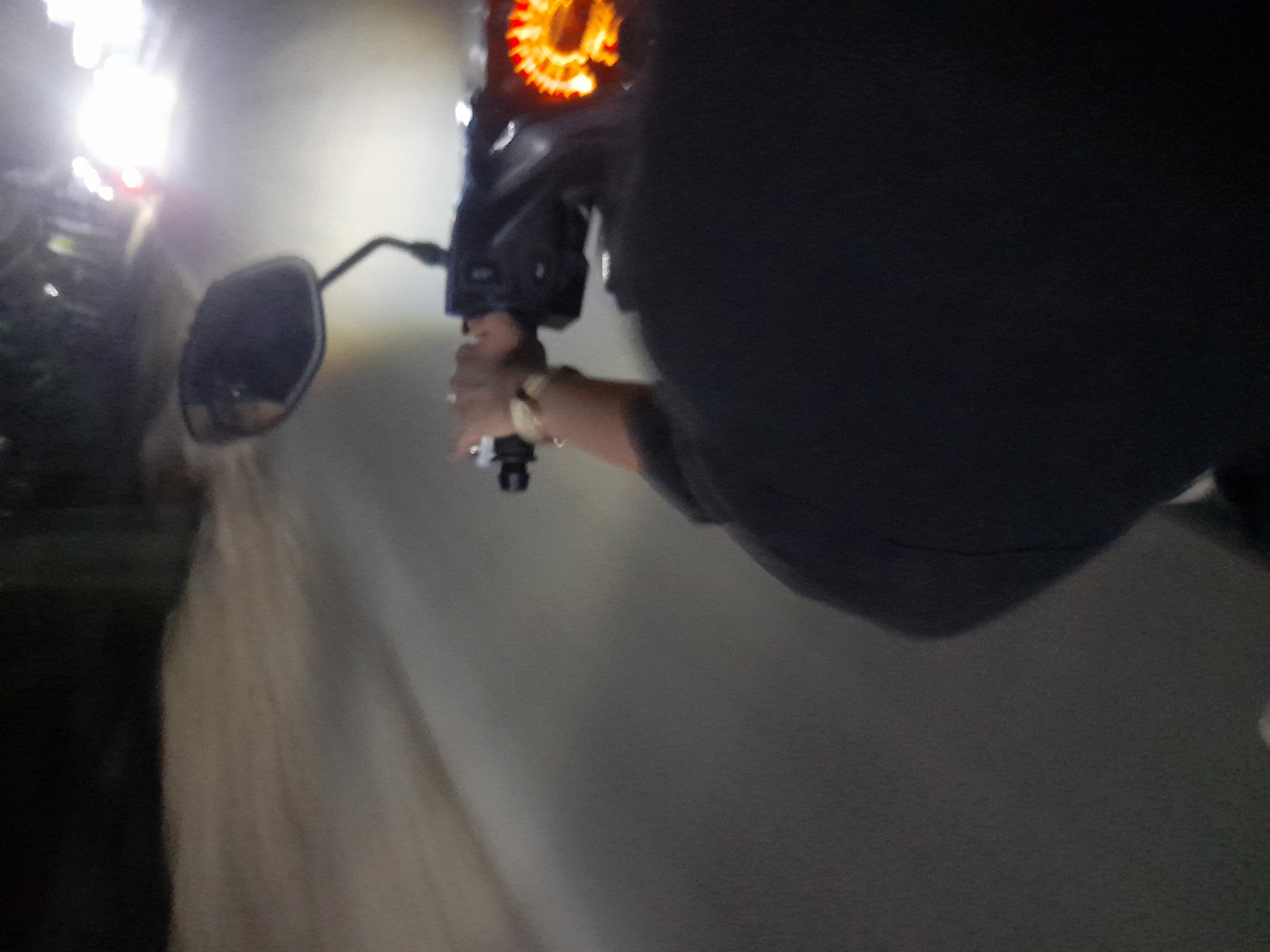 The image captures a nighttime scene from the perspective of someone riding a motorcycle in Jetaksari, Indonesia. The foreground is dominated by the shoulder and back of the rider, wearing a dark hoodie. Their right hand is visible on the handlebars, adorned with a gold watch. A portion of the motorcycle's dashboard with illuminated indicators is also visible. The mirror of the motorcycle reflects a bright light, likely from another vehicle. The road ahead is paved and appears wet, reflecting the ambient light from streetlights and other vehicles. Trees and foliage are faintly visible on the sides of the road, indicating a semi-urban or rural setting. The overall atmosphere is dark, with artificial lights creating streaks and blurs due to motion.