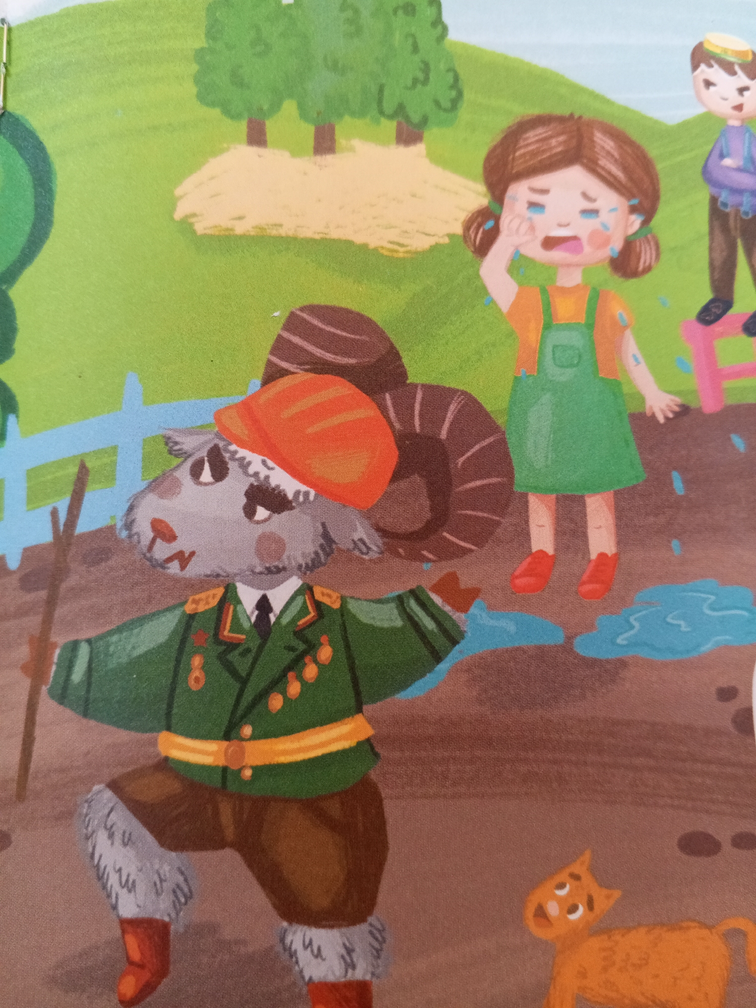 The image depicts an illustration of a pastoral scene with cartoon characters. In the foreground, a ram wearing a green military-style jacket with medals, brown pants, and an orange cap is standing and holding a wooden stick. Next to the ram, a young girl with brown pigtails is crying, with tears streaming down her face and on her green overalls and orange shirt. Behind her, a boy in a purple shirt and brown pants stands on a pink stool, looking on. The ground is brown with puddles of blue water scattered around, and a small orange cat is visible in the lower right corner. In the background, there are green trees and a yellow dirt path. The scene does not provide any specific visual cues or landmarks to suggest a location in Chistopol, Russia.