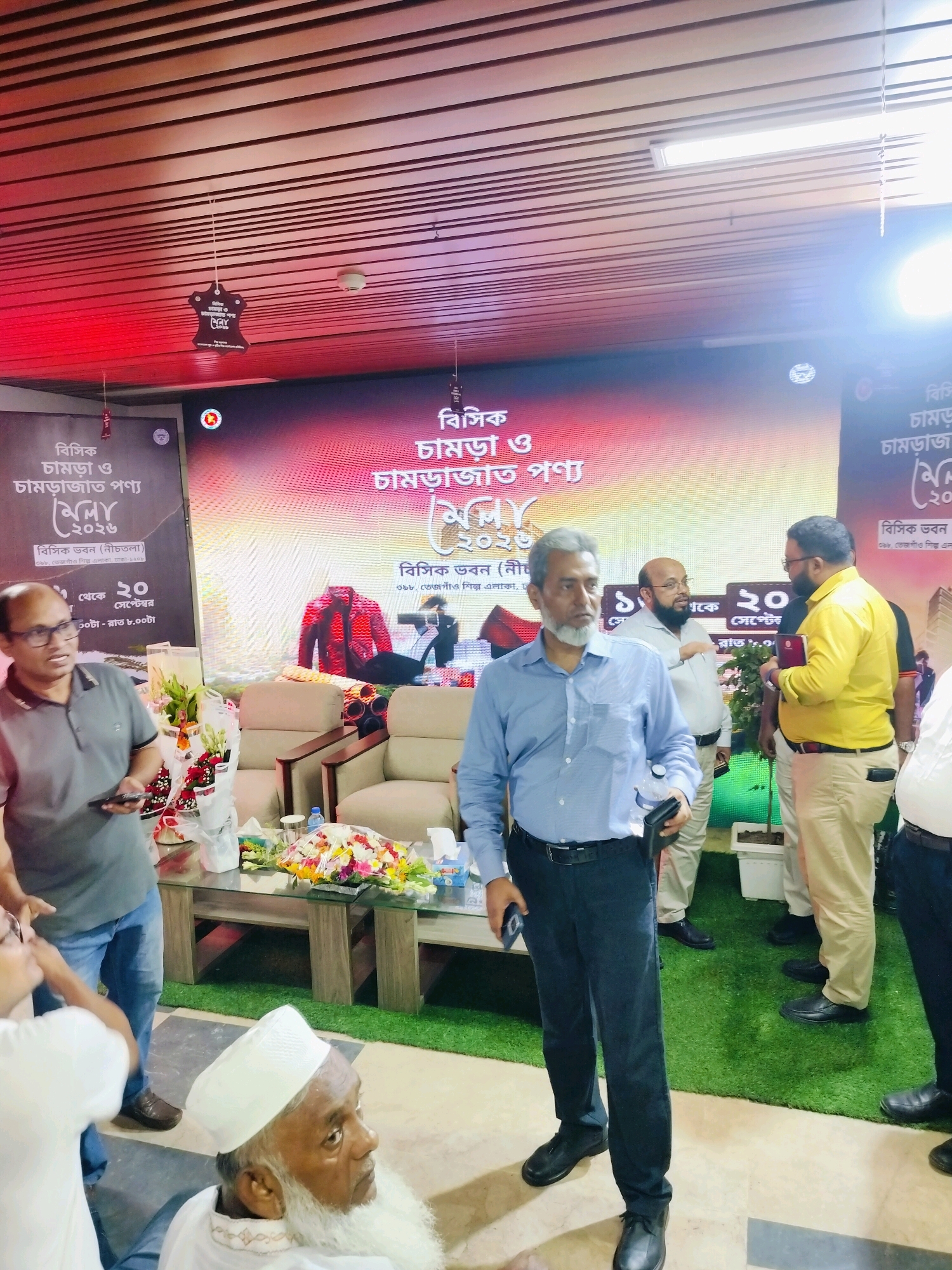 In this wide shot taken indoors, likely at an event in Dhaka, Bangladesh, a group of men stand near a large digital display that features Bengali text and graphics. The display shows images of clothing and text that appears to be promotional for an event. The ceiling is made of dark wooden slats with red lighting. In the foreground, a man in a blue shirt and dark pants is holding a phone and a wallet. To his right, another man in a yellow shirt and light-colored pants is holding a book and talking to a man in a grey shirt. Further back, a man with a beard and glasses in a light-colored shirt is also engaged in conversation. A table with flower arrangements and bottles of water is in the middle ground, with chairs visible behind it. An elderly man with a white beard and a white cap is seated in the lower right portion of the frame. The floor is covered in part by green artificial turf.