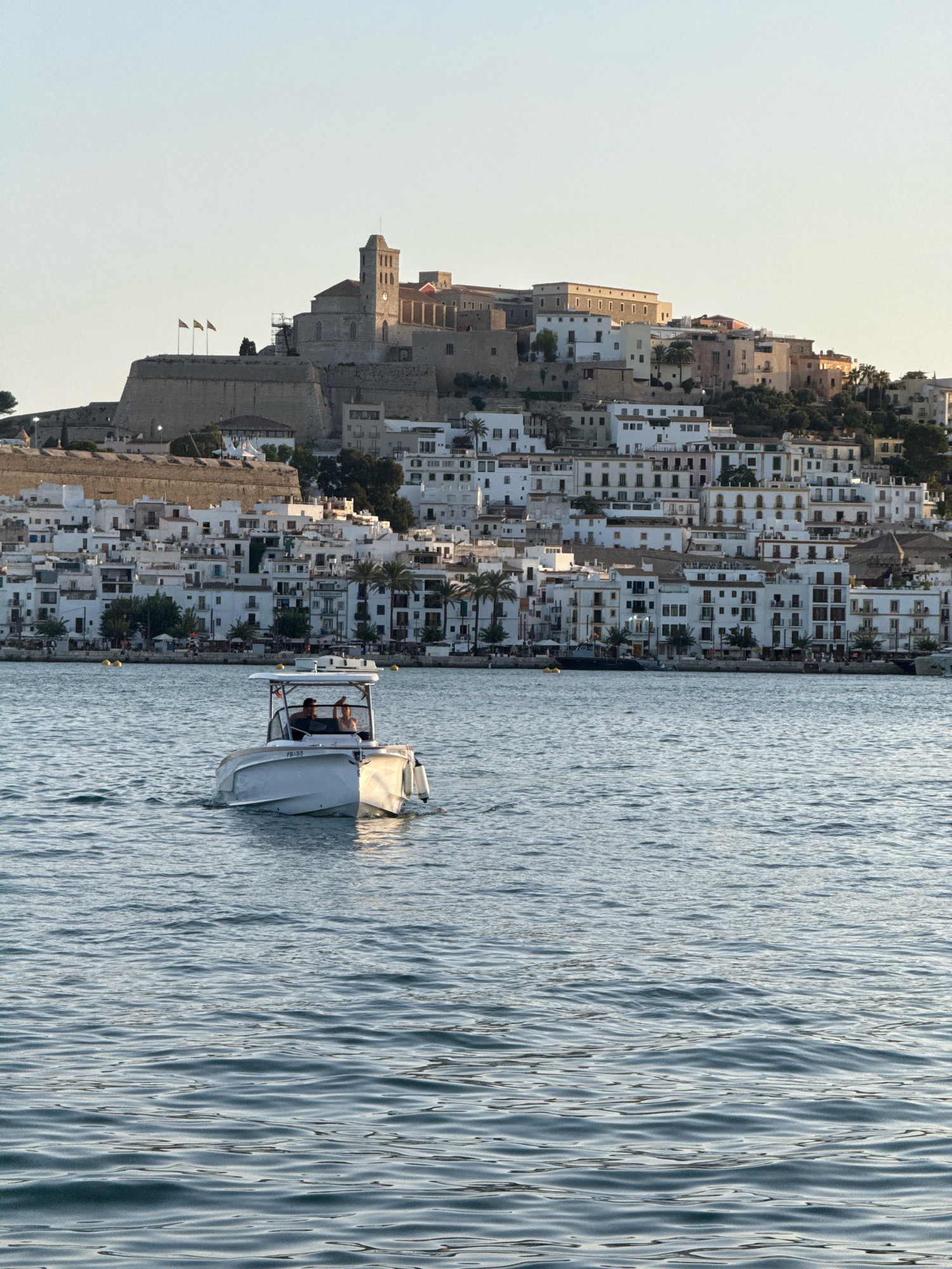 The image depicts a daytime scene of the city of Eivissa, Spain, with a focus on the historic Dalt Vila area overlooking the water. In the foreground, a white motorboat with a canopy is navigating the blue water. Two individuals are visible in the boat, one steering and the other appearing to wave. The boat has a license plate that reads "FM-1-15-S". In the middle ground, a cluster of white buildings forms the densely packed cityscape of Eivissa, cascading down the hillside. Above the city, the imposing stone walls and structures of Dalt Vila, a UNESCO World Heritage site, are prominent. A church tower with a clock is a notable feature of the fortified citadel. Three flags are visible flying from a battlement on the left side of Dalt Vila. The sky is clear and pale blue, suggesting fair weather. The overall impression is one of a Mediterranean coastal city bathed in natural light.