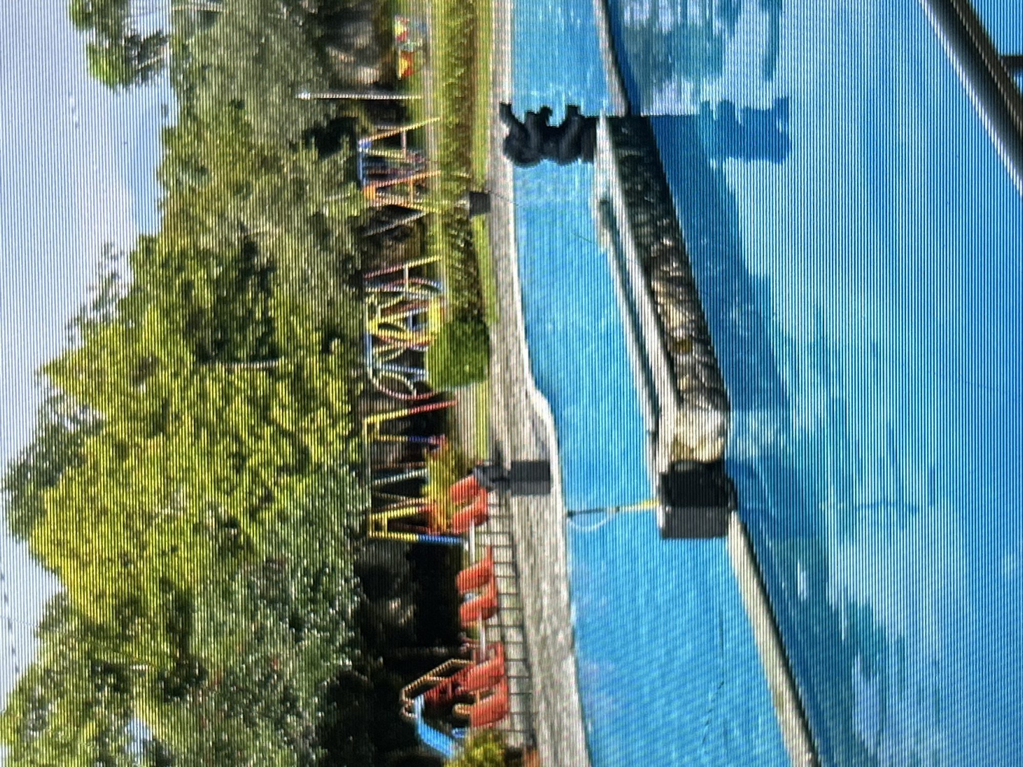 The image depicts a swimming pool area with a playground in the background, located in Depok, Indonesia. The swimming pool is large and filled with bright blue water. A stone-like structure with a decorative elephant statue sits at the edge of the pool. Several lounge chairs are arranged on a tiled area next to the pool, some of which are empty. In the background, a playground with colorful swings and a slide can be seen, surrounded by lush green trees and vegetation. The sky is visible above the trees, appearing light blue.