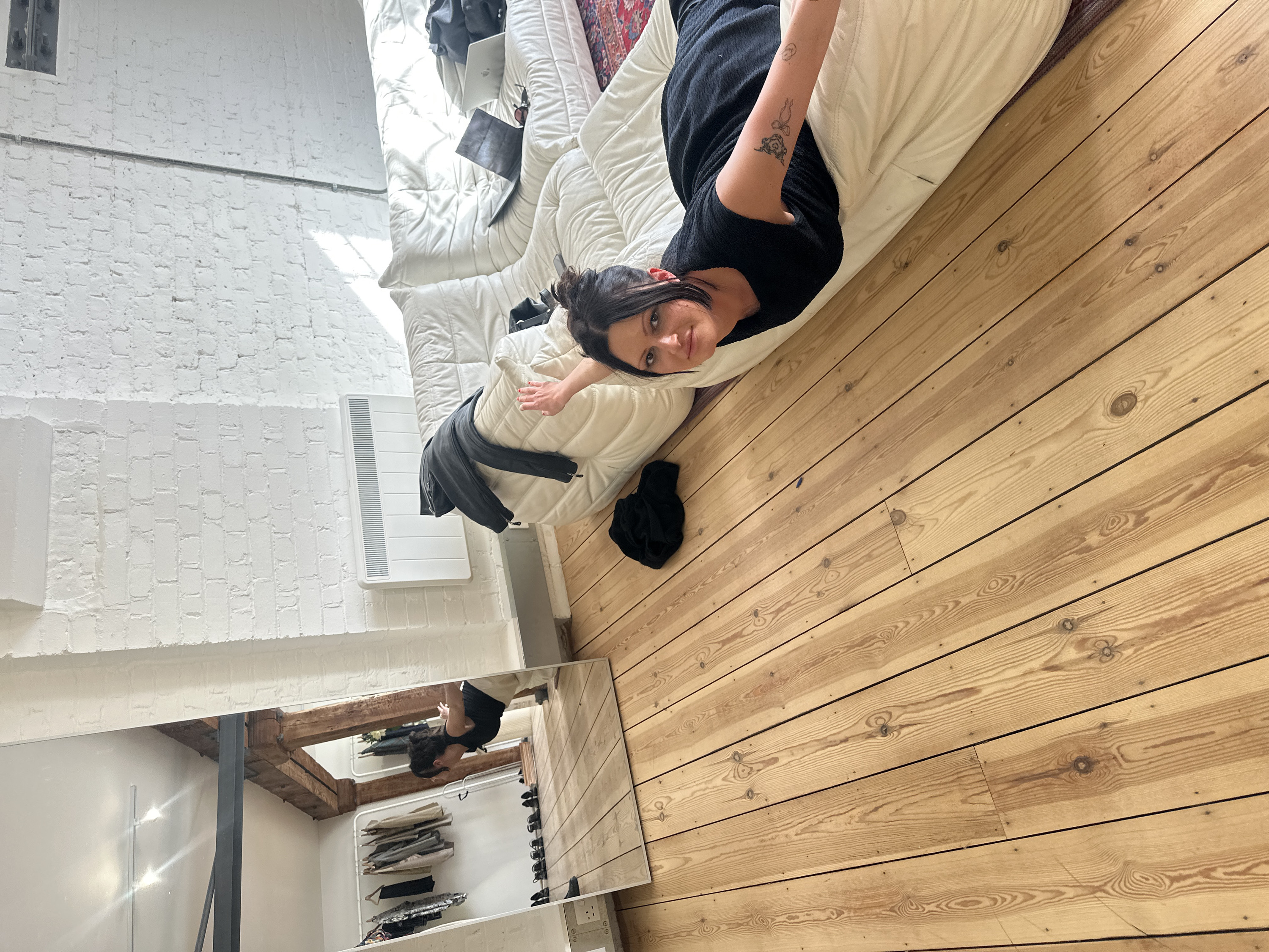The image depicts the interior of a renovated urban loft or studio in London, United Kingdom. The space features white-painted exposed brick walls and a light-toned wooden plank floor. Exposed wooden structural beams are visible, with an additional metal crossbeam reflected in a large, floor-standing mirror on the left side of the frame. The ceiling, partially visible in the reflection, has white track lighting. A white wall-mounted electric heater is installed on the right wall.
A female individual with dark hair tied back and bangs is lying prone on a large, segmented white sofa, positioned towards the right side of the frame. She is wearing a dark, possibly black, short-sleeved top or dress and is looking directly at the camera. Her left arm is extended outwards, parallel to the floor, displaying multiple line-art tattoos on her forearm. Her right hand, with red nail polish, is also extended outwards.
On the white sofa, a black jacket is draped over a section. Further along the sofa, two open laptops are visible: one dark, the other a silver Apple MacBook. A pair of dark sunglasses rests between them. A patterned rug, possibly Persian-style, is partially visible beneath the sofa. A piece of black clothing is crumpled on the wooden floor next to the sofa.
The large, rectangular mirror on the left reflects another view of the woman, who appears to be standing or leaning slightly forward, wearing a black top and light-colored trousers, with both arms extended horizontally. The reflection also reveals a white metal clothing rack holding various garments, several pairs of black shoes arranged on the floor beneath the rack, and a white UK-standard double power outlet on the lower wall.