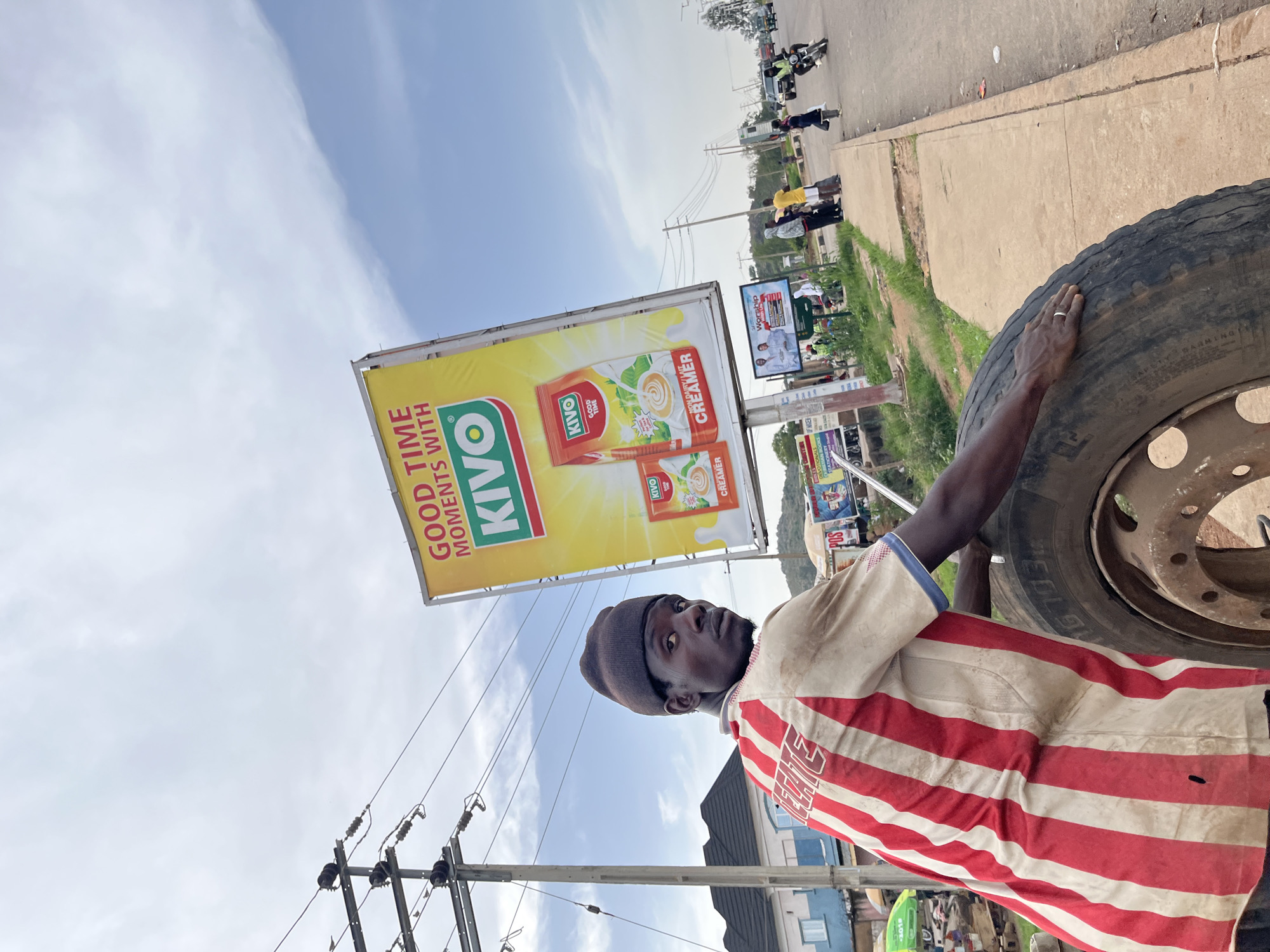 A man stands next to a large tire, his right hand resting on its surface. He is wearing a red and white striped t-shirt and a dark beanie. In the background, a large billboard advertises "KIVO GOOD TIME MOMENTS WITH KIVO CREAMER". Other smaller billboards and poles with electrical wires are visible. The scene appears to be outdoors, possibly on a roadside in Nigeria, given the location context. The sky is cloudy, suggesting it might be daytime. The man's expression is neutral. The overall scene suggests an urban or semi-urban environment with commercial activity.