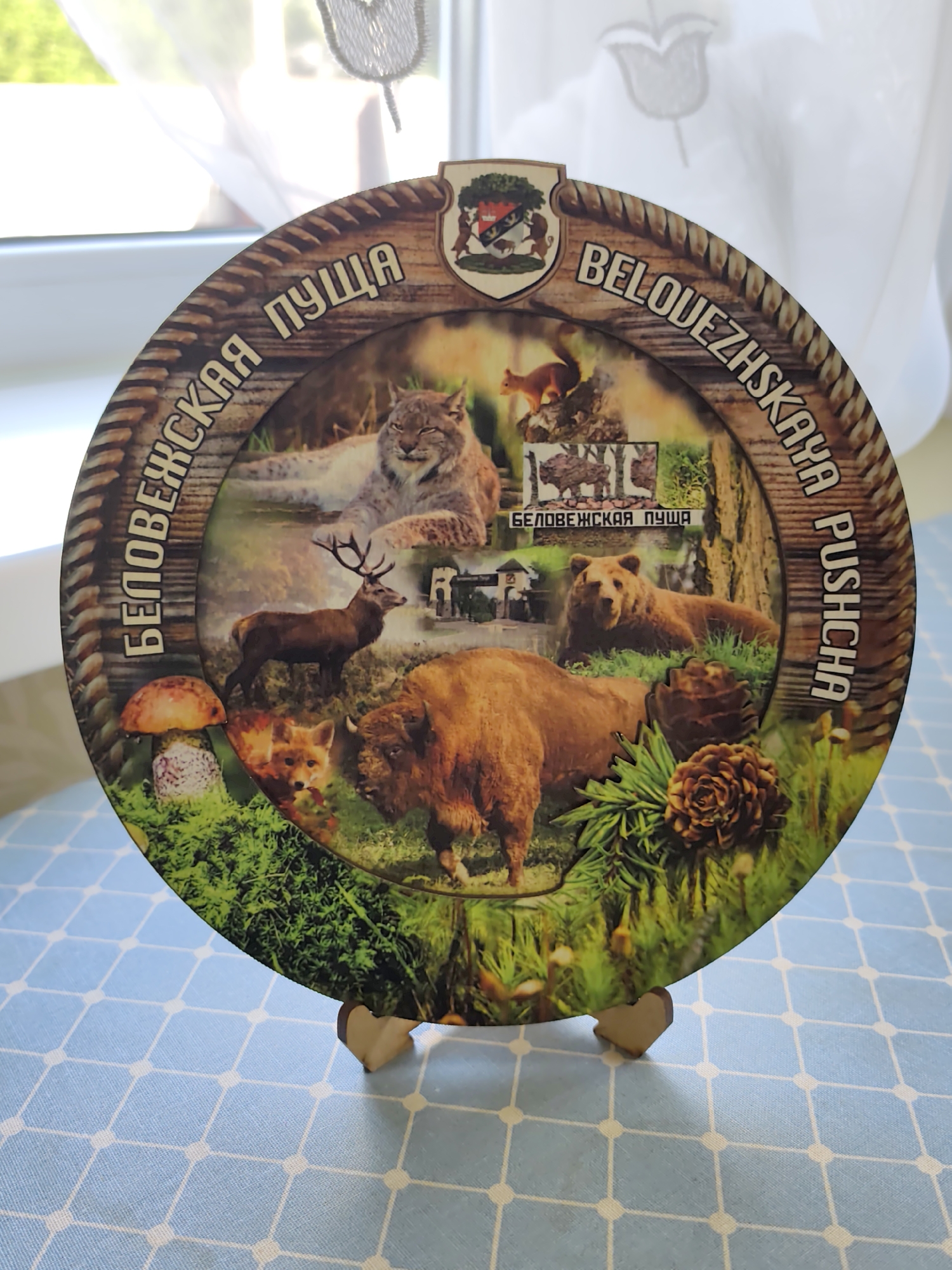 This is a close-up, eye-level, slightly angled shot of a round decorative plate featuring a collage of wildlife and text. The plate is resting on a light blue and white checkered tablecloth. The plate's border has a rope-like texture and is decorated with images of various forest animals, including a lynx, a deer, a fox, a bison, and a bear. There are also images of mushrooms and pine cones. In the center of the plate, there are smaller images of a bison and a building, with the text "БЕЛОВЕЖСКАЯ ПУЩА" written below them. The outer edge of the plate has text in a curved arrangement, reading "БЕЛОВЕЖСКАЯ ПУЩА" on the left side and "BELOUEZH SKAYA PUSCHCHA" on the right side. A small shield with a coat of arms is visible at the top center of the plate. The background is blurred and appears to be an indoor setting with a window and sheer curtains. The plate is supported by two small, light-colored wooden stands. The overall impression is that of a souvenir or decorative item. The text "БЕЛОВЕЖСКАЯ ПУЩА" refers to the Białowieża Forest, a protected area that straddles the border between Poland and Belarus, near the village of Бараўлянскі сельскі Савет.