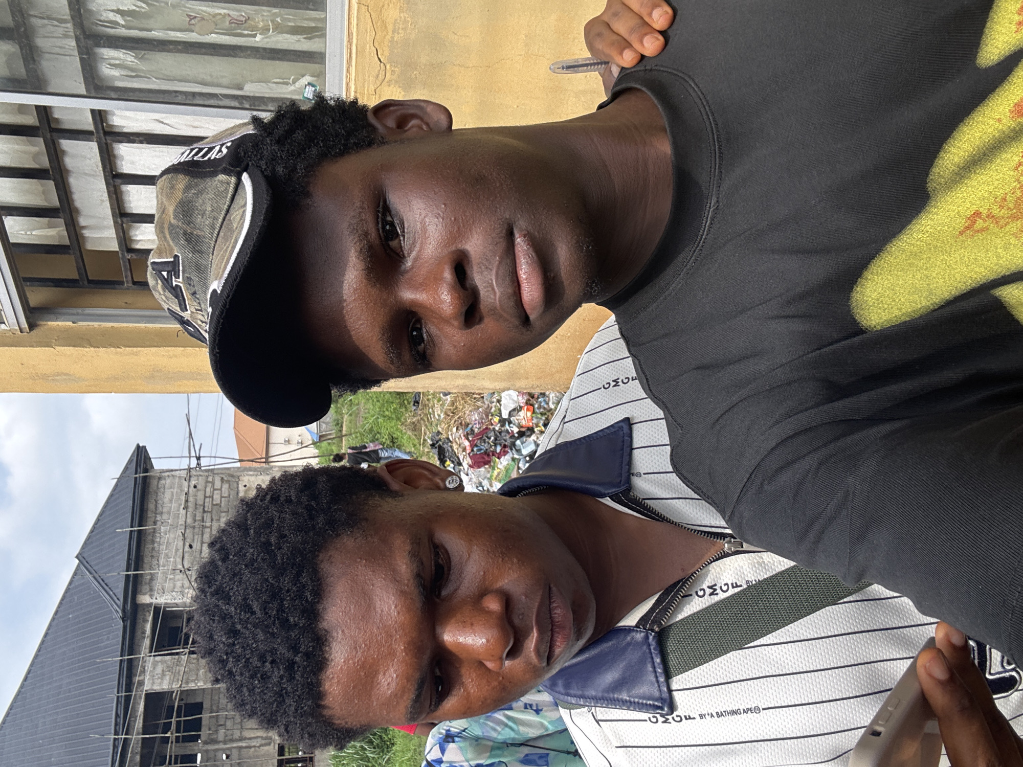 Two young Black males are posing for a selfie in front of a yellow building in what appears to be a semi-urban area. The male on the left is wearing a white shirt with thin vertical blue stripes and a dark blue collar, with a green strap across his chest. He has a large, circular, silver earring in his left ear. The male on the right is wearing a black t-shirt with a yellow graphic design. He is wearing a camouflage baseball cap with a black brim. The male on the right has his arm around the shoulder of the male on the left. In the background, there is a building under construction with scaffolding, and a pile of debris is visible to the right of the male on the left. The sky is overcast. This scene is located in the city of Ihiagwa, Nigeria.