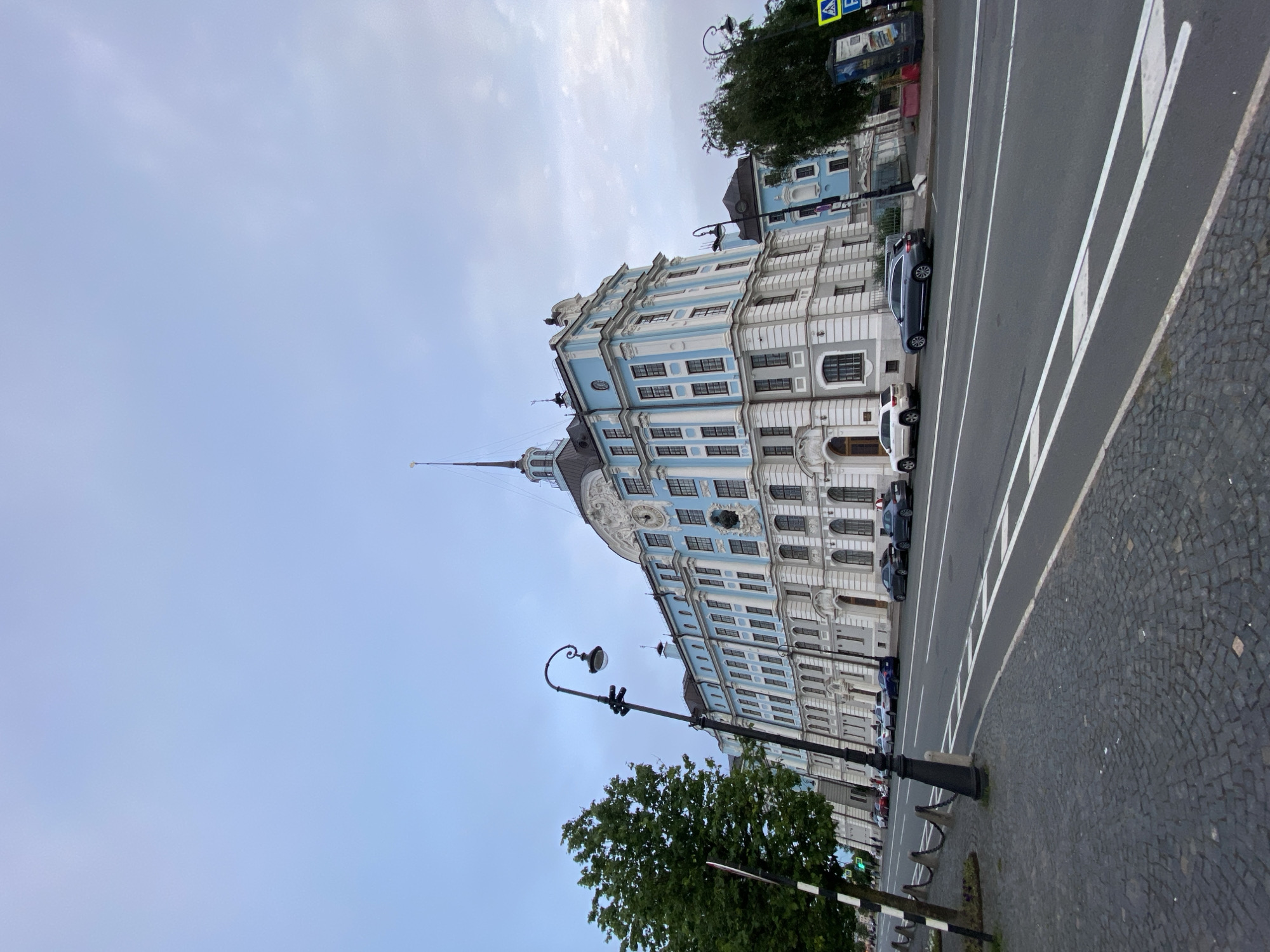 The image depicts a wide, gray street in Saint Petersburg, Russia, lined with cobblestone pavement on the right and a large, ornate, light blue and white building on the left. The building features multiple stories with numerous windows, decorative moldings, and a prominent clock on its facade. A tall, slender spire rises from the roof of the building.
Several cars are parked along the street. A classic-style lamppost stands near the building, its curved arm holding a single light fixture. Trees with dense green foliage are visible in the foreground and background. The sky is overcast and pale, suggesting a cloudy day. The orientation of the image is vertical, as if taken looking up at the building from street level. The scene appears to be set during the daytime. There is a traffic light visible in the lower right corner and a pedestrian crossing painted on the road.