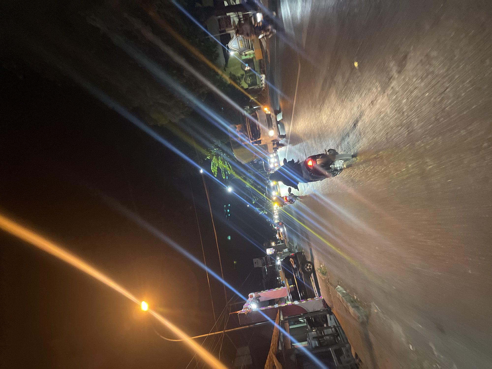 The image depicts a street scene at night in Kalinyamatan, Indonesia. The perspective is from a moving vehicle, likely a motorcycle, looking forward and slightly to the right. A street light with a strong yellow glow is prominently visible in the upper left quadrant, casting a distinct beam across the dark sky. The road surface appears wet and reflects the various lights present, creating streaks of color.
In the foreground, the rear of a motorcycle is visible, with its brake light illuminated. The rider is wearing dark clothing. Further ahead on the road, there are other vehicles, including a white truck with its headlights on and a darker SUV or van parked on the left side of the road. Several other motorcycles and cars are visible in the middle ground, their headlights and taillights creating bright points and trails of light.
Along the left side of the street, structures with awnings are visible, suggesting a commercial or residential area. One building has colorful lights decorating its facade. Trees are present on the right side of the road, their silhouettes dark against the night sky. Traffic signals are also visible in the distance. The overall impression is of a busy street at night, with vehicles and streetlights contributing to a dynamic visual environment.