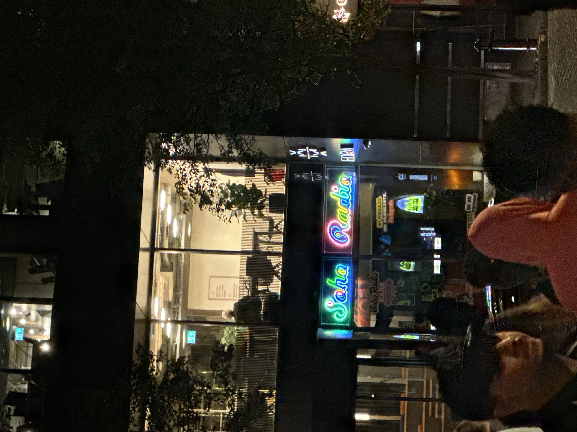 The image captures a nighttime scene in London, United Kingdom, featuring the Soho Radio storefront. Illuminated neon signs spelling out "Soho Radio" in vibrant colors dominate the view. Reflections and interior details are visible through the large glass windows, including silhouettes of people inside, light fixtures, and furniture. Outside, trees frame the scene, adding depth to the composition. Passersby are also visible in the foreground. The overall atmosphere is urban and vibrant, typical of a bustling city environment at night.
