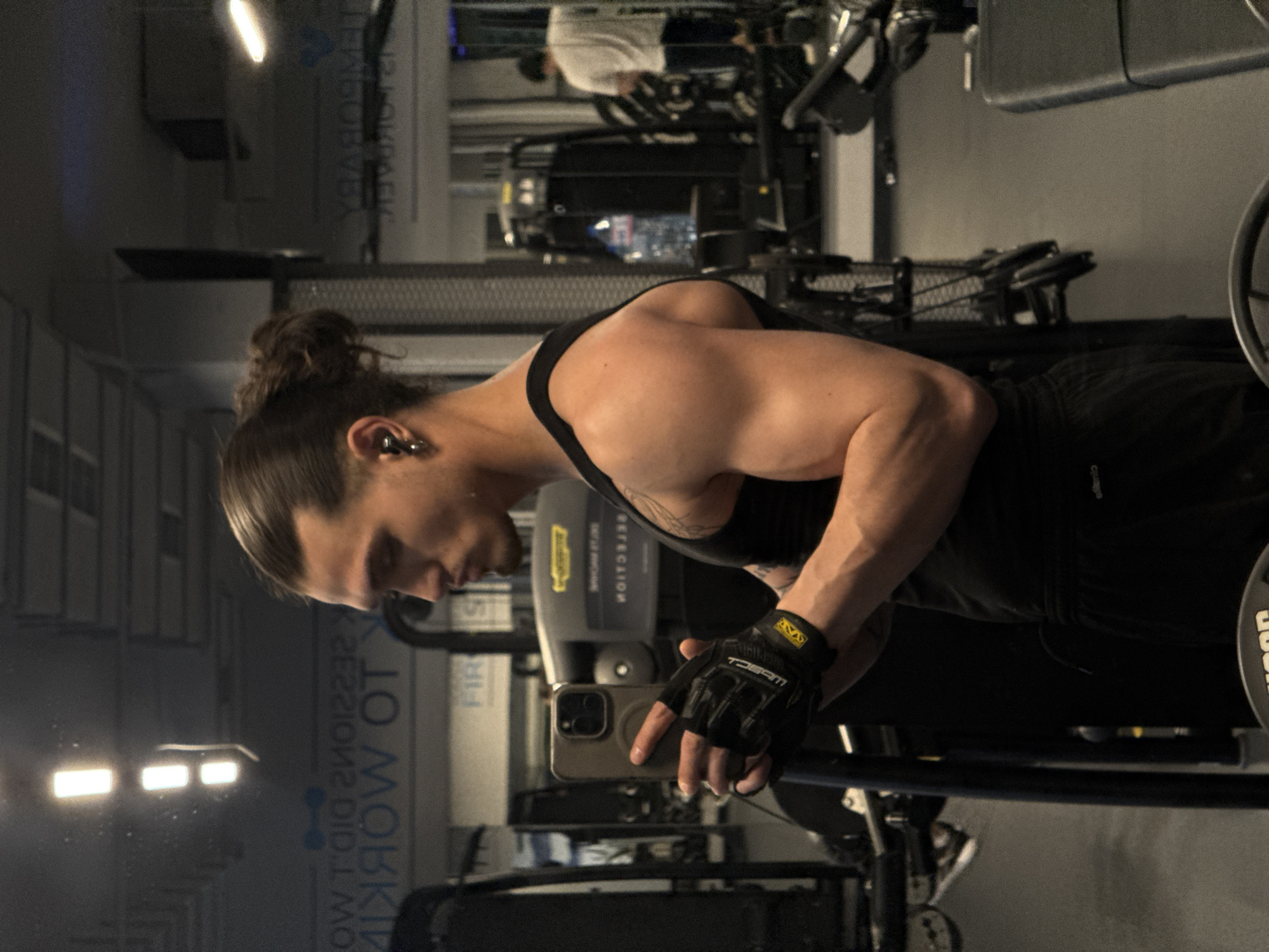 A male individual, appearing to be in his twenties or thirties with dark hair tied in a bun, is taking a mirror selfie in a gym located in Sofia, Bulgaria. He is wearing a black tank top, black athletic shorts, and black fingerless weightlifting gloves, with visible tattoos on his left arm/shoulder and an earbud and earring in his right ear. He holds a dark-colored smartphone with a circular grip accessory in his gloved left hand, angled to capture his reflection. The gym environment, seen in reflection, features various exercise machines, including one with a reversed "TECHNOGYM" logo, and a clear plastic water bottle on a machine. Reflected text on the wall includes phrases such as "SESSIONS DID WORK," "TO WORK," and "TEMPORARY," alongside a blue dumbbell icon. Overhead fluorescent lighting is visible as bright rectangular reflections. In the far background, another person in a white t-shirt is visible from behind, interacting with gym equipment. The flooring is dark, and the walls are light grey.
