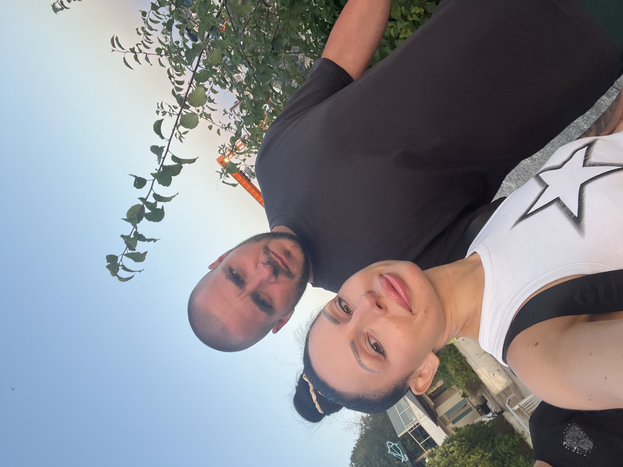 A selfie is taken outdoors during the day, with a clear sky overhead and trees and buildings in the background. The foreground is dominated by two people. On the left, a woman is looking directly at the camera with a slight smile. She has dark hair pulled back into a bun and is wearing a white tank top with a black star design. On the right, a man is also looking towards the camera, with a neutral expression. He has a shaved head and is wearing a black t-shirt. The background shows some greenery, a building with balconies, and a bright orange structure, possibly part of a crane or construction equipment. The setting appears to be a public area or walkway in the city of Primorsko, Bulgaria, given the architectural style of the building and the presence of vegetation typical of coastal regions.
