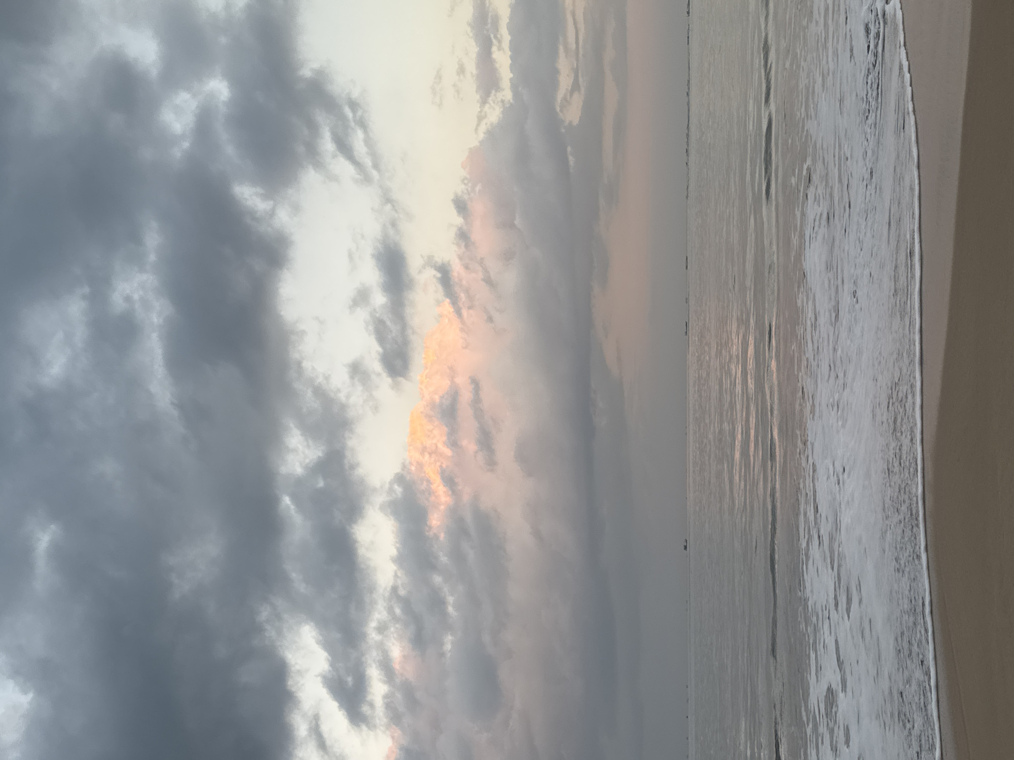 A serene beach scene in Kuta, Indonesia, features a calm ocean with gentle waves lapping onto a sandy shore. The sky is filled with dramatic, heavy clouds, predominantly gray, but with a striking band of warm, soft pink and orange hues breaking through the overcast. A few distant boats are visible on the horizon, scattered across the vast expanse of the sea, suggesting a peaceful and quiet atmosphere. The sand in the foreground is smooth and wet from the receding tide, reflecting the muted colors of the sky.