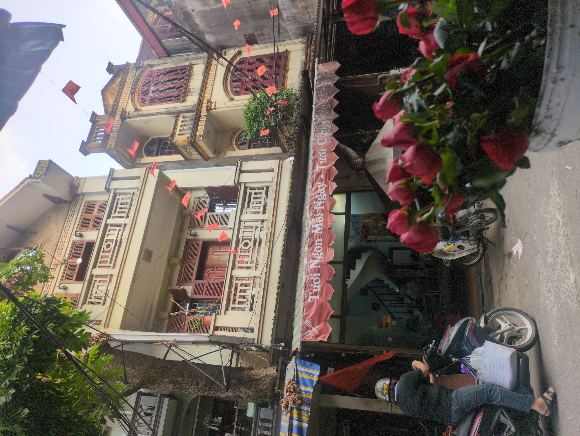 A street view in Hà Nội, Vietnam, shows a row of traditional, narrow houses with balconies and red-shuttered windows. Red triangular flags are strung across the street between the buildings, and a large tree with dense foliage partially obscures the upper floors on the left.
In the foreground, a person wearing a black shirt, blue jeans, and a helmet is seated on a black and silver motorbike. The motorbike has a large white woven basket attached to the front. The person is looking towards the left, away from the viewer. To the right, another motorbike, mostly white, is parked.
A red awning stretches across the front of one of the buildings, displaying Vietnamese text that reads "Tươi Ngon Mỗi Ngày - Tươi Cũ." Below the awning, the interior of a shop is visible, with a spiral staircase leading upwards.
In the extreme foreground, a cluster of red roses in a white bucket is out of focus, with some petals scattered on the ground. The street appears to be paved with asphalt. The sky is overcast and bright.