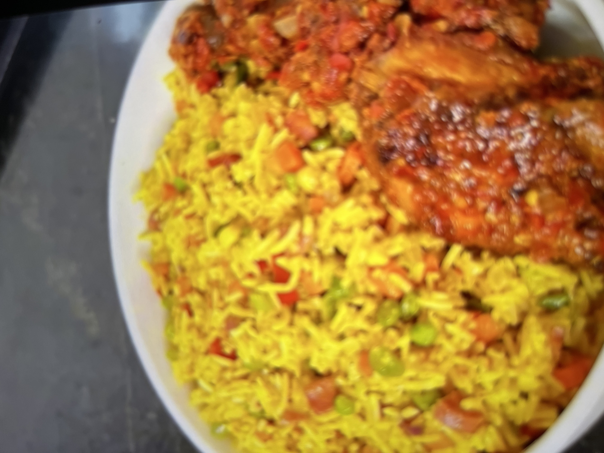 A close-up, slightly out-of-focus shot shows a white bowl filled with food. The bowl contains a mound of yellow rice mixed with chopped vegetables, including red bell peppers, green peas, and carrots. To the right of the rice, there are several pieces of meat coated in a reddish-brown sauce. The food is presented in what appears to be a restaurant or home setting, with a dark, textured surface visible in the background, possibly a countertop or table. The lighting is warm, highlighting the colors of the dish. There are no people or specific landmarks of Abuja, Nigeria, visible in the image.