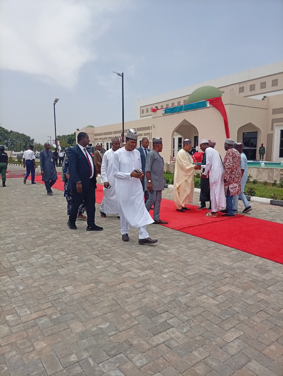 This outdoor image captures a formal gathering of men on a paved pathway leading to a prominent building in Unguwan Sanusi, Nigeria. A red carpet extends along the right side of the path, signifying an official event.
In the foreground, a man in a white kaftan and a traditional cap, holding a mobile phone, walks towards the left. Behind him and slightly to his left, another man in a dark suit and red tie is also walking. Numerous other men are visible, most dressed in various forms of traditional Nigerian attire, including long kaftans and caps, while some wear Western suits.
On the red carpet to the right, a group of men is actively engaging in greetings, with several individuals shaking hands. One man in a light-colored kaftan is seen extending his hand in a handshake to another man in white, surrounded by other men in patterned and plain traditional garments. Security personnel in dark uniforms are also present further back in the scene.
The background features a modern, light-colored building characterized by arched entrances, rectangular windows, and a distinctive green dome on its roof. A red and green banner or fabric is draped near the dome. The architecture suggests a public, institutional, or ceremonial building, possibly with Islamic influences. The ground is covered with grey interlocking paver blocks, and tall lampposts line the pathway. The sky is bright with visible clouds, indicating a daytime setting. No specific text is discernible in the image.