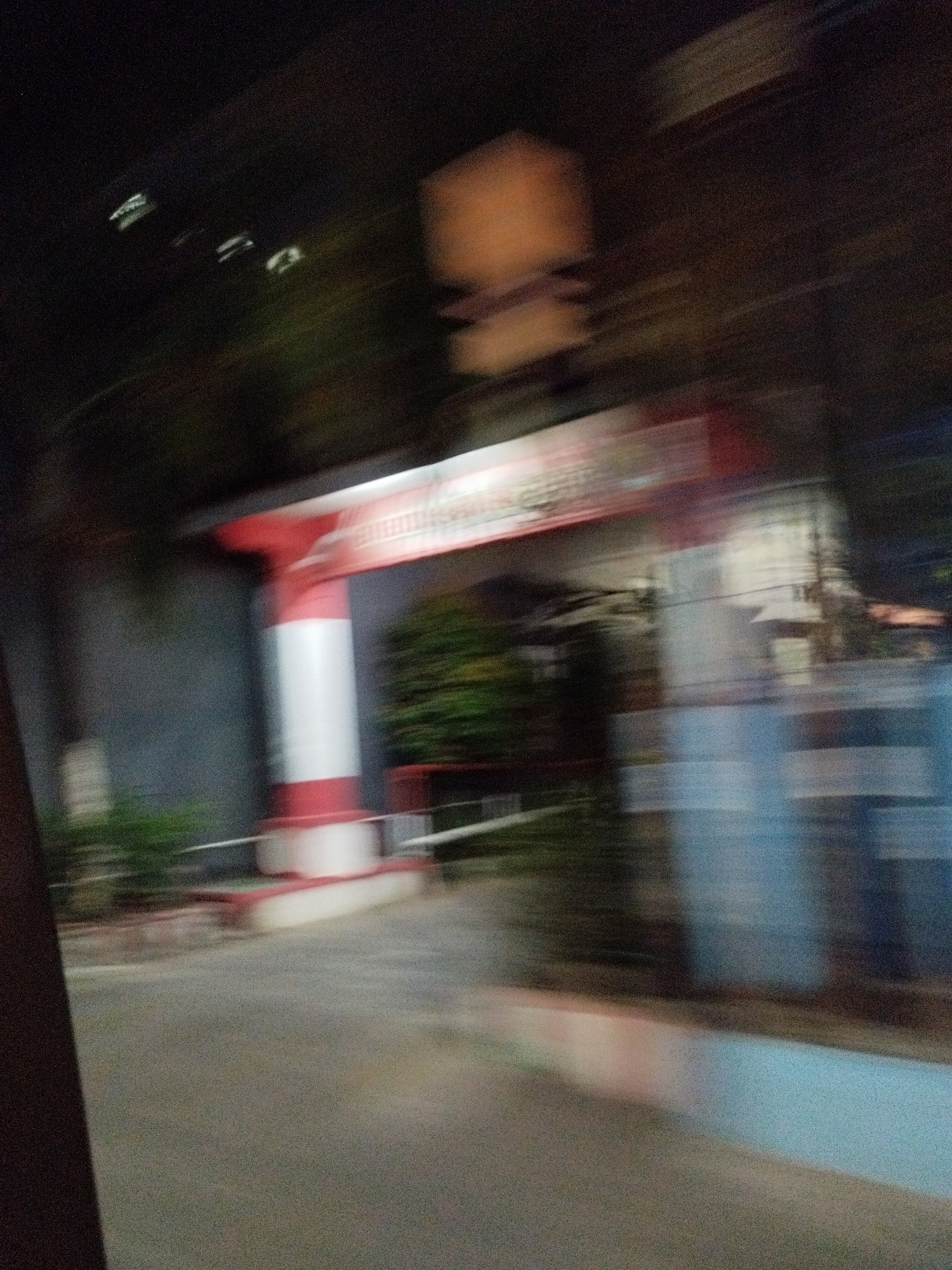 The image depicts a blurry night scene, likely captured from a moving vehicle, in Surabaya, Indonesia. The most prominent feature is a red and white striped archway, possibly an entrance to a building or establishment. The arch has text on it, but it is illegible due to the motion blur. To the left of the arch is a grey wall and some greenery. To the right, there are blurred structures and lights, indicating an urban environment. The ground in the foreground appears to be a paved road. The overall impression is one of a fleeting glimpse of a location at night.