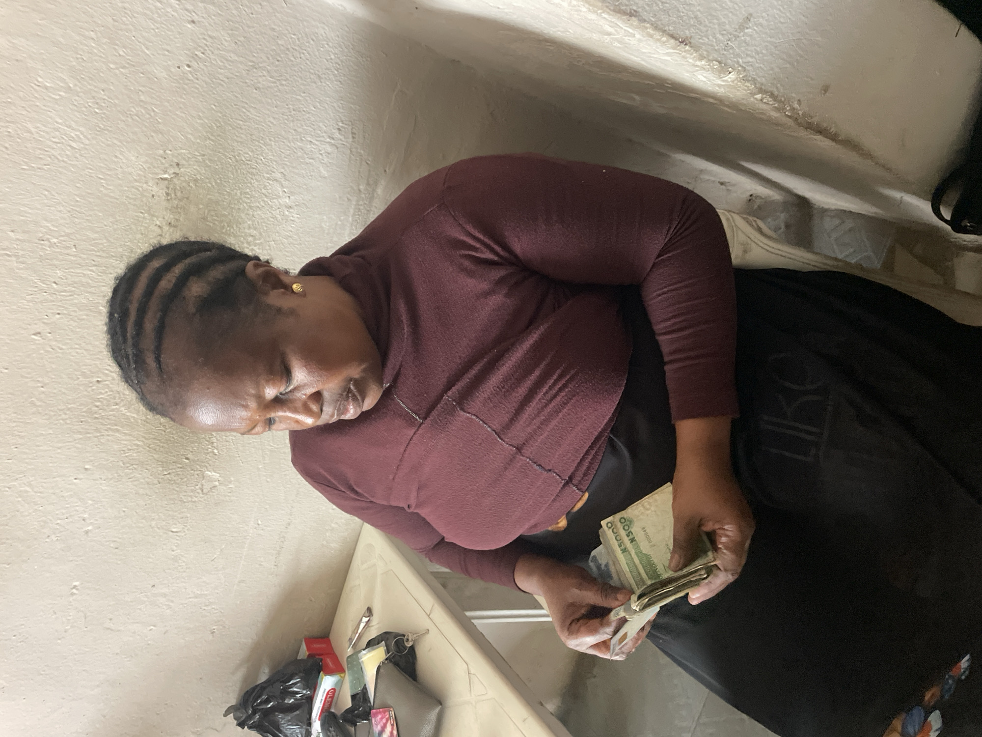 A woman with dark skin and braided hair is seated, holding a wad of Nigerian 5000 naira banknotes. She is wearing a dark maroon turtleneck top and a black skirt. The woman is looking down at the money in her hands, which she appears to be counting or organizing. She has a gold earring in her visible ear. She is seated next to a light-colored table on which a black plastic bag, a red box with a white label, and a grey wallet with keys are placed. The background consists of a textured, off-white wall and a partially visible chair. The scene is indoors, likely in a residential or modest commercial setting in Jos, Nigeria.