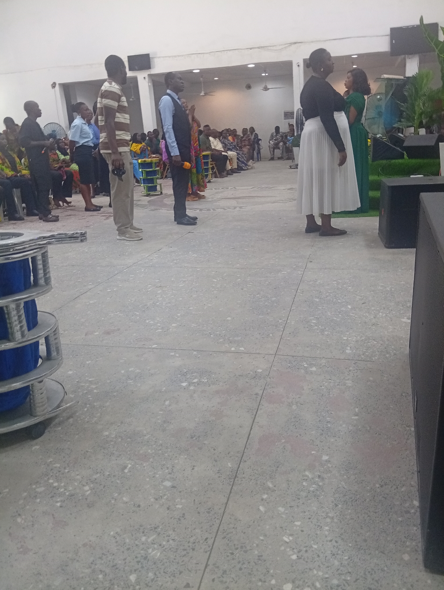 This is a medium-shot, eye-level, full-color photograph of a group of people gathered indoors. The setting appears to be a community hall or event space, with a polished concrete floor. In the foreground, on the left, is a tall, cylindrical object with metallic silver banding and blue material coiled inside. To the right of this object, several people are standing in a line, facing away from the camera and towards the background.
The individuals in the foreground are dressed in various attire. One man in the center wears a striped t-shirt and khaki pants. To his right, another man is dressed in a vest and collared shirt, holding a microphone. Further to the right, a woman in a long white skirt and dark top is visible, and next to her, another woman in a green dress.
In the middle ground, a crowd of people are seated and standing. The background reveals a white wall with ceiling lights and ceiling fans. There are also speakers visible on the right side of the image. The overall atmosphere suggests an organized gathering or service. The location is identified as Kason, Nigeria.