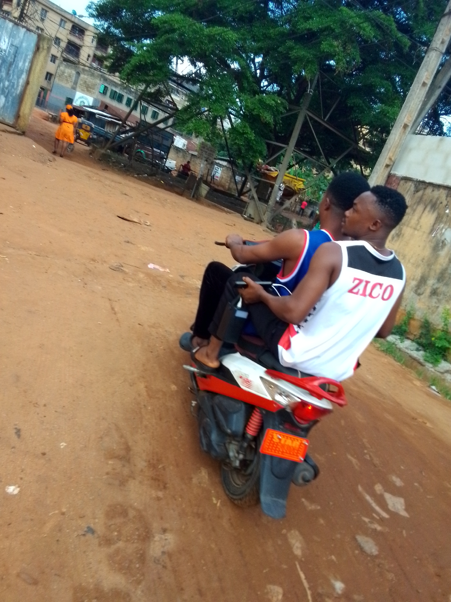 Two young men on a red and white scooter drive along a reddish-brown dirt road. The passenger in the back wears a white tank top with the name "ZICO" printed on the back in red letters. The driver wears a blue basketball-style jersey. Both are wearing black pants. The driver wears sandals.
In the background, a woman in an orange dress walks away from the camera. Behind her are buildings and what appears to be a yellow auto rickshaw or "keke" parked. A large tree looms in the upper-right.
The dirt road suggests a less developed area within a city, possibly Onitsha, Nigeria. The presence of buildings, power lines, and vehicles indicate an urban area. The open-air environment, clothing style, and type of transportation are consistent with a West African context.