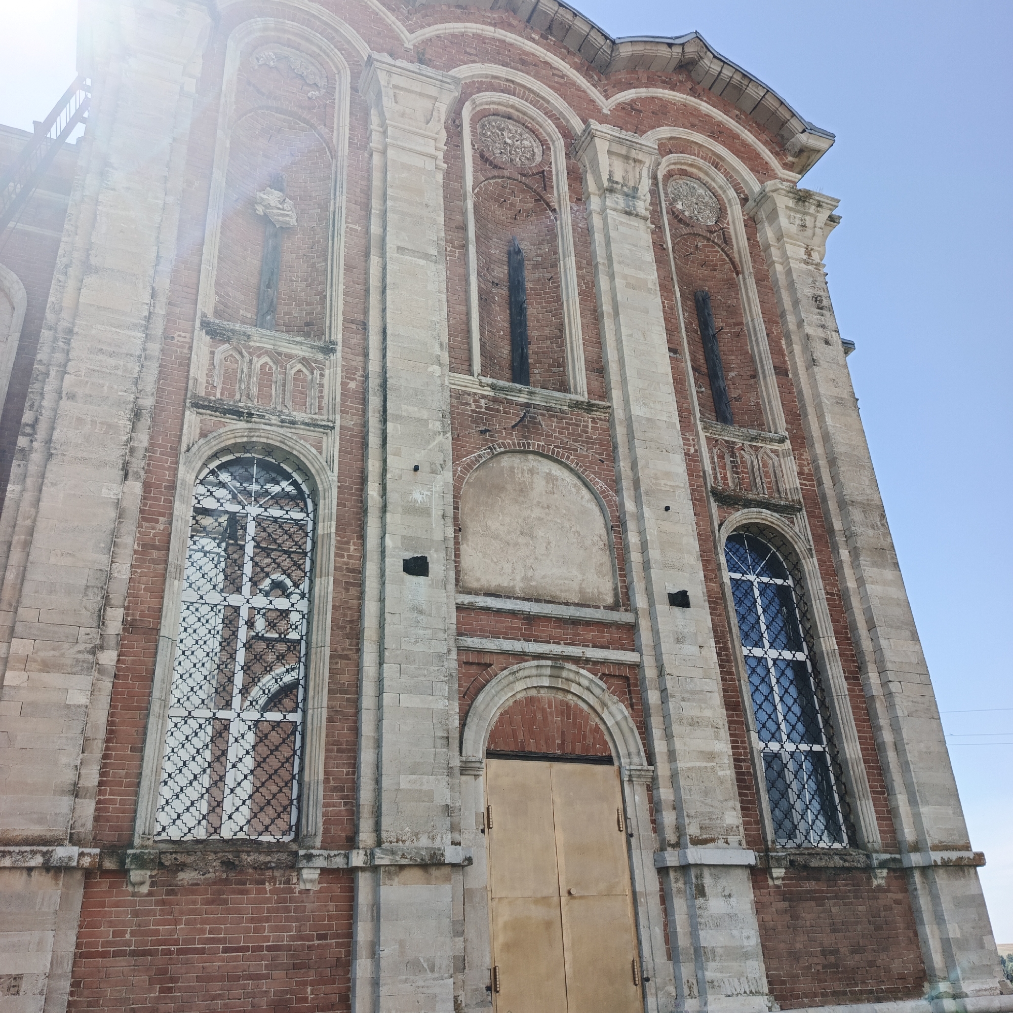 The image shows the facade of a red brick building with stone accents and arched windows. The building appears to be an old church or a similar public structure, likely in the village of Tugutdemir, Russia. The facade features decorative stone columns and arched niches, some containing remnants of structures. Two large arched windows with intricate metal grates are visible, as well as a double door with a golden finish at the base. A bright sun glare is present in the upper left portion of the image, casting a strong light on the building and sky. The sky is clear and blue. The overall condition of the building suggests it is an historical landmark, possibly in a state of disrepair or undergoing restoration, given the exposed brickwork and weathered stone.