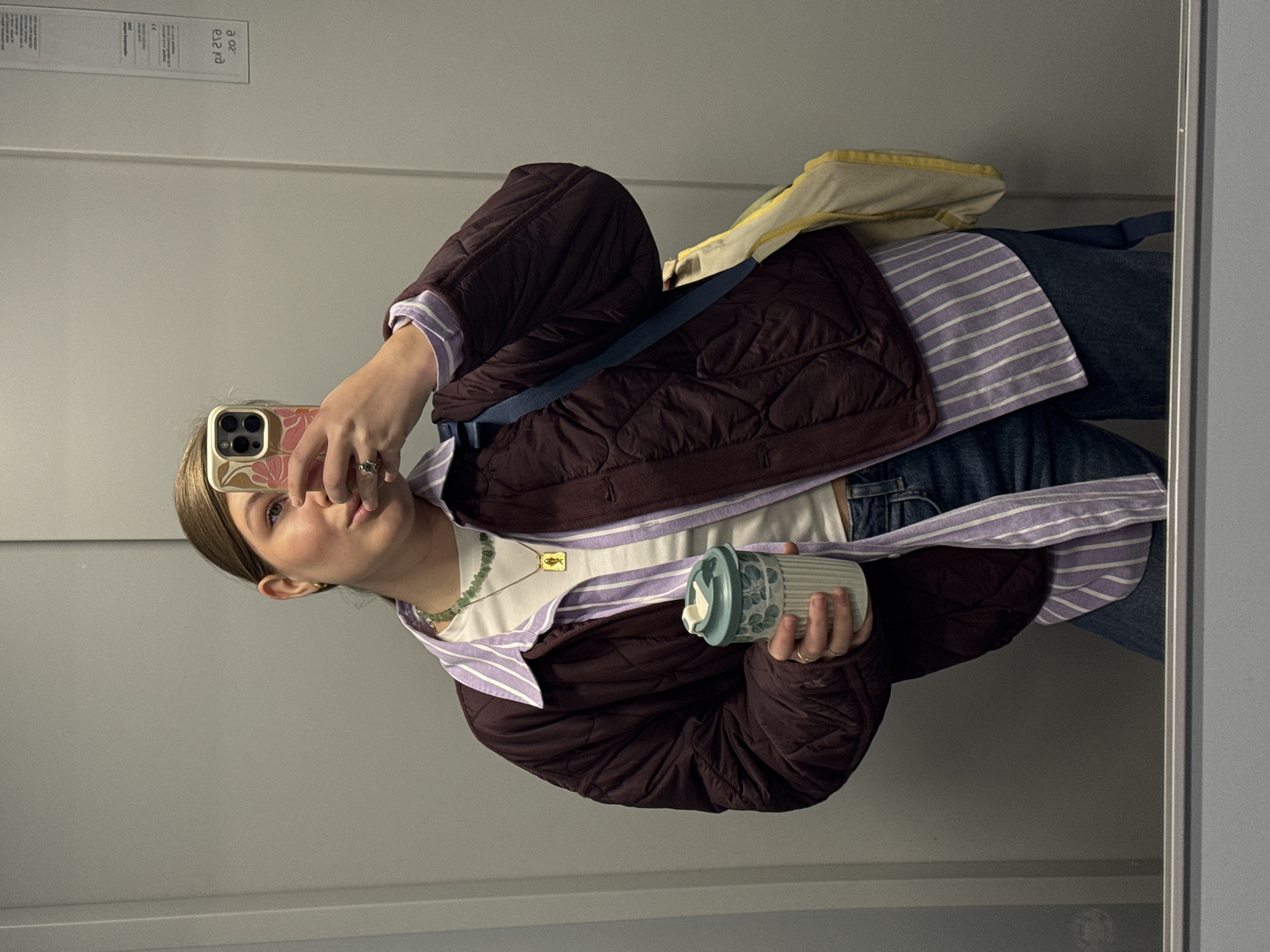 A person is standing in front of a mirror, holding a coffee cup in one hand and a cell phone in the other, apparently taking a selfie. The person is wearing a brown quilted jacket over a white t-shirt and a purple striped shirt, with jeans. The image appears to be taken indoors, possibly in an elevator. The specific location within Vozdovac Urban Municipality, Serbia, cannot be determined from the image.