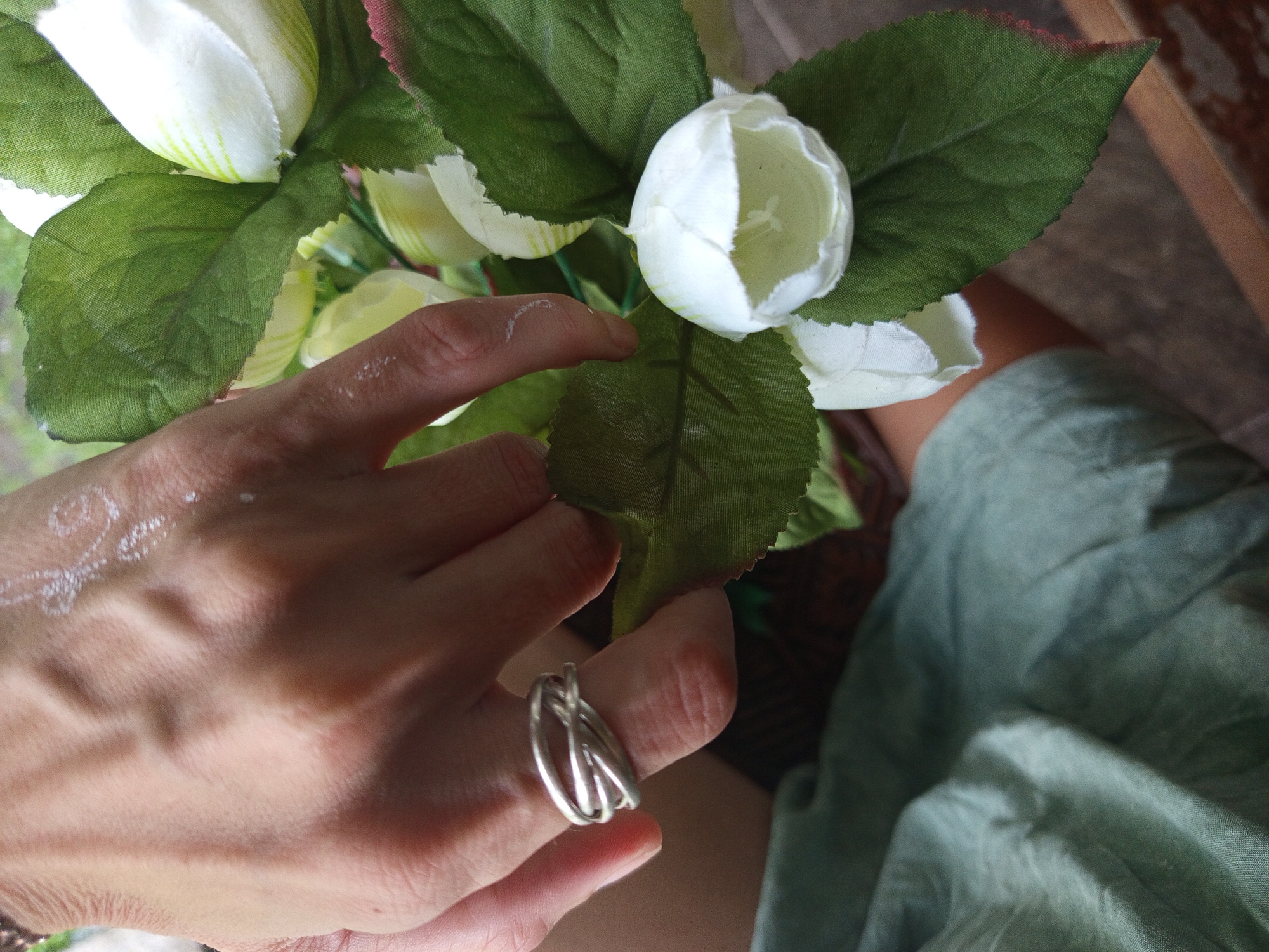 A close-up, high-angle photograph captures a person's hand gently interacting with an arrangement of white and green flowers. The hand, with light skin tone, features faint white decorative lettering on its back, which appears to spell "Love". A distinctive silver-toned, multi-banded ring is worn on the ring finger. The flowers are predominantly white, tulip-like buds, with some showing hints of pale green stripes, accompanied by vibrant green, possibly artificial, leaves. The hand is shown lightly touching one of these leaves. In the lower right background, crumpled light green fabric, suggestive of clothing like a skirt or sarong, is visible, alongside a dark, blurred surface that might be wood. The scene is illuminated by natural daylight, indicating a daytime setting. The overall impression is one of calm and appreciation for the flowers, located in North Kuta, Indonesia.