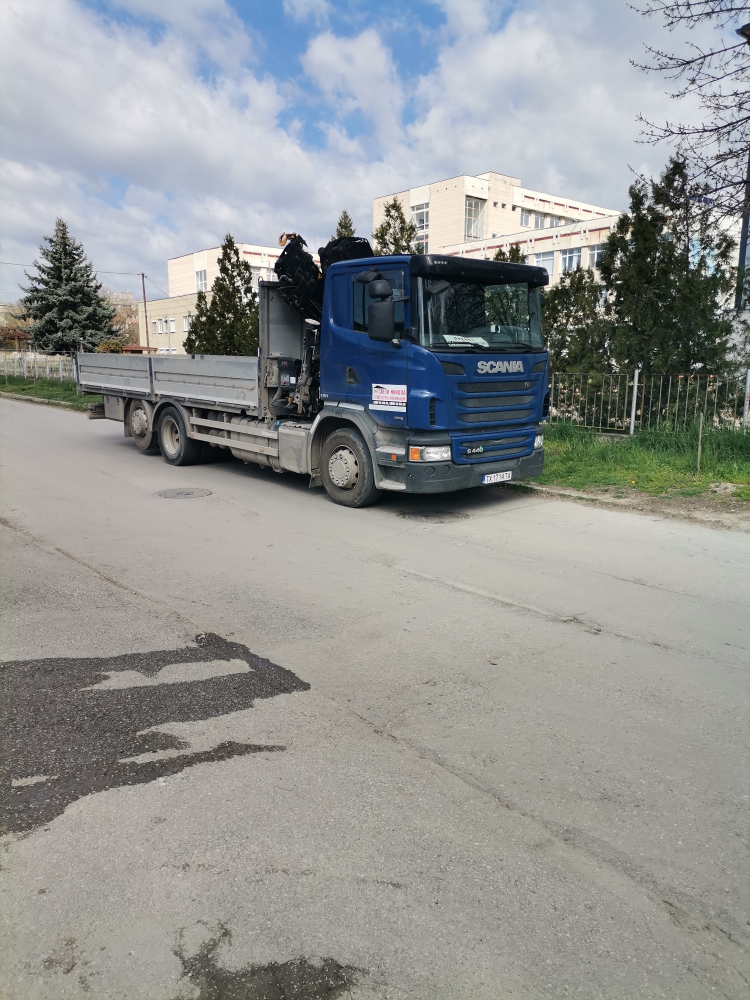 The image depicts a blue Scania G 440 truck parked on an asphalt road in a likely urban setting. The truck has a flatbed and a crane mounted behind the cab. A building with multiple stories and windows stands in the background, surrounded by evergreen trees and a metal fence. The sky above is partly cloudy. The license plate of the truck is TX 171 TA. The location is most likely in Dobrich, Bulgaria, as indicated by the truck's license plate and the Cyrillic text on the truck's door. There are shadows and patches on the road surface in the foreground. No people are visible in the image.