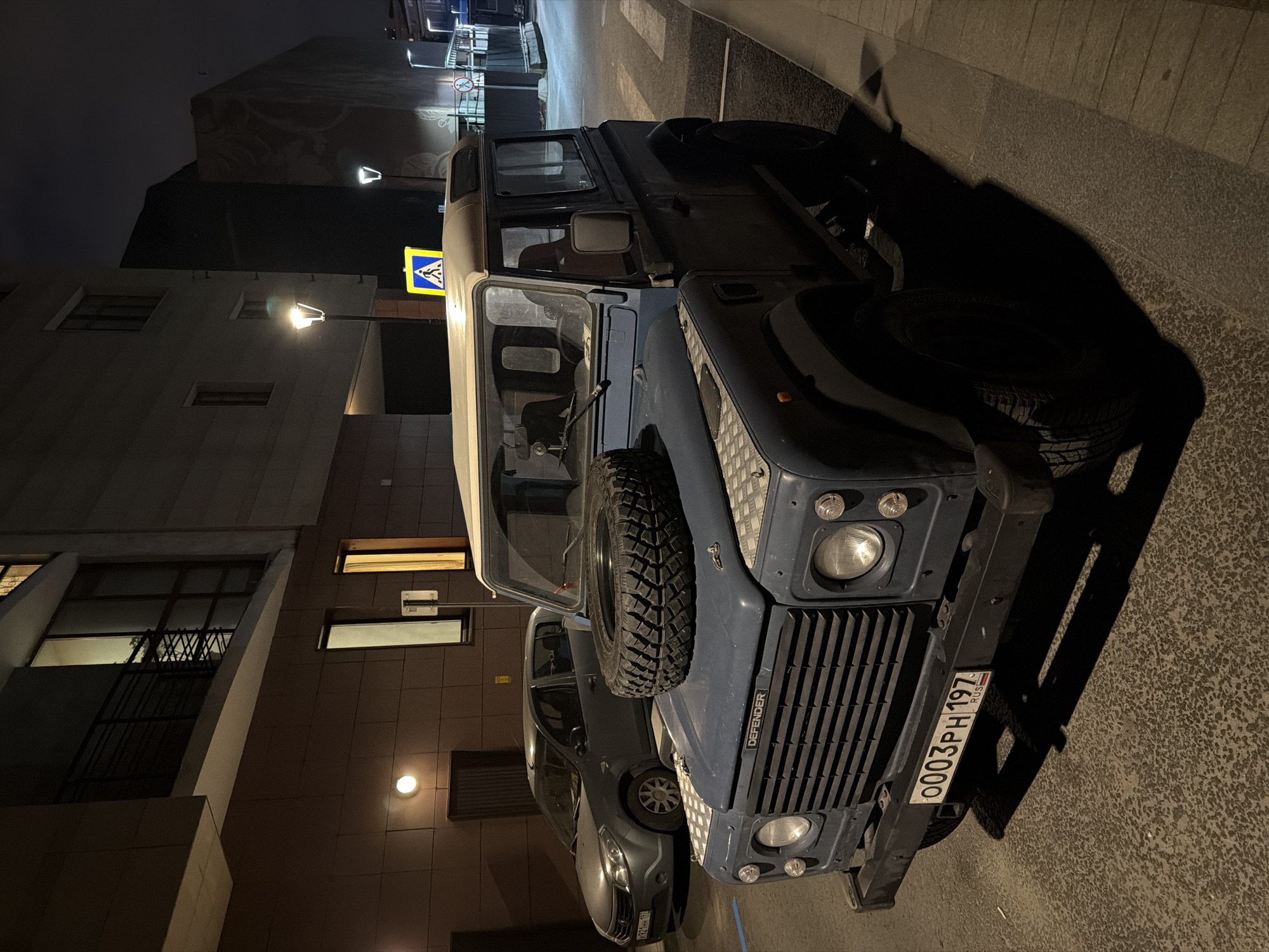 The image shows a blue Land Rover Defender parked on a city street at night. The Defender is positioned in the foreground, with its front grille and hood visible. A spare tire is mounted on the hood. To the left of the Defender, a grey sedan is parked. The background features a modern building with illuminated windows and balconies. Streetlights are visible, casting light on the scene. A pedestrian crossing is marked on the road in front of the Defender. A yellow pedestrian crossing sign is mounted on a pole in the background. The license plate of the Defender reads "0003PH 197 RUS". The scene appears to be in an urban environment, likely a residential or commercial area in Moscow, Russia. There are no people visible in the image.